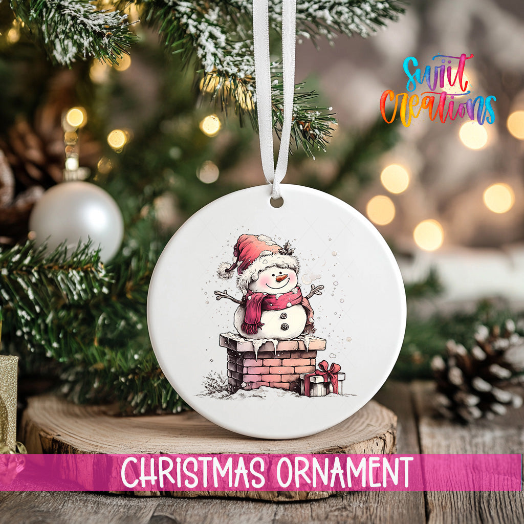 A white Christmas ornament featuring a snowman is hanging from a ribbon, with a blurred background of a decorated Christmas tree and other ornaments.