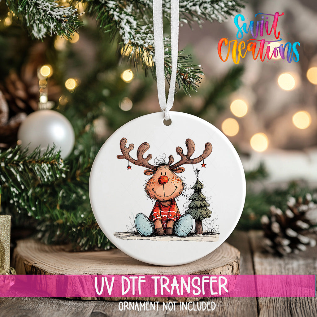A white ceramic ornament with a cartoon reindeer and Christmas tree is hanging on a Christmas tree.
