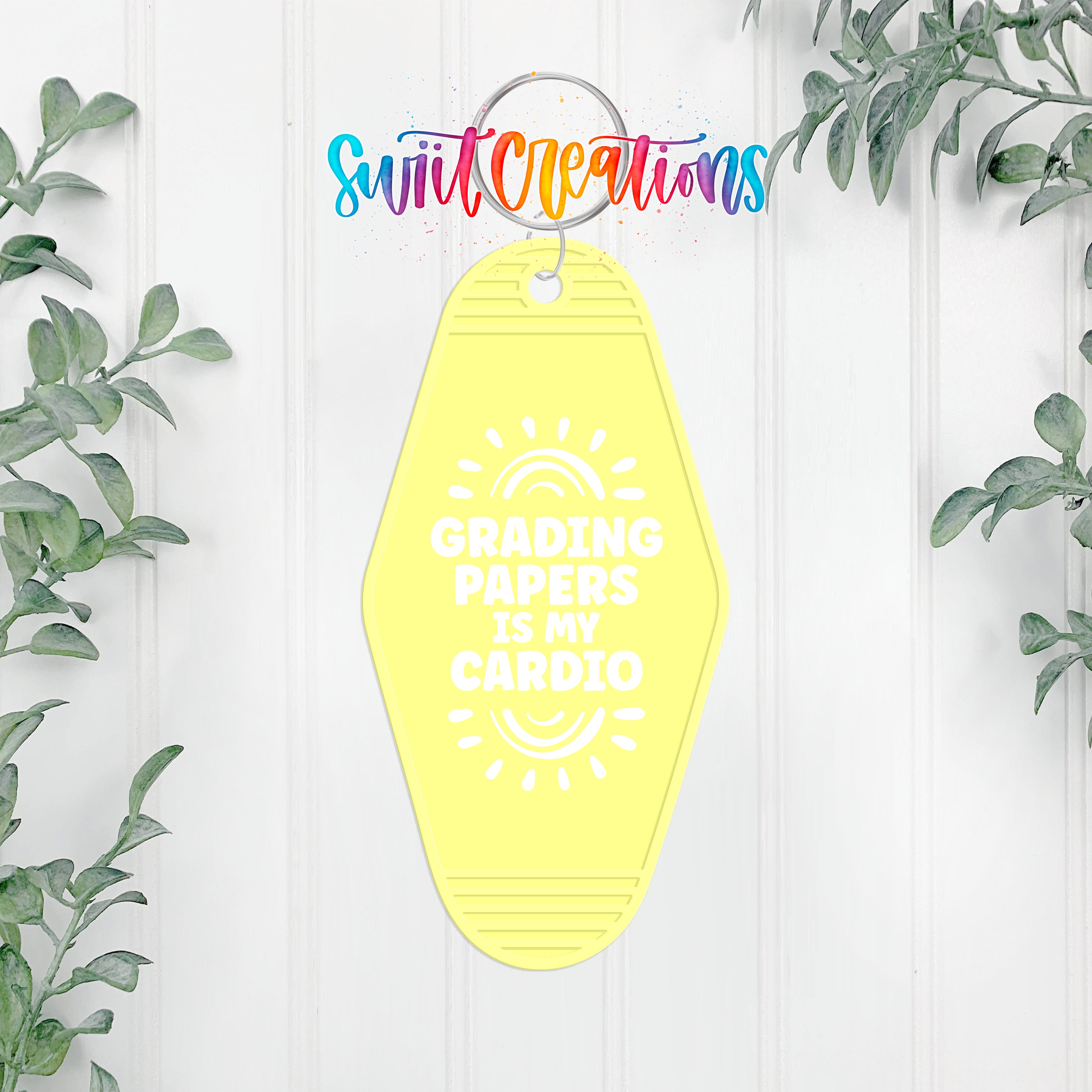 A yellow keychain with the text "GRADING PAPERS IS MY CARDIO" printed on it, hanging on a white wall with greenery in the background.