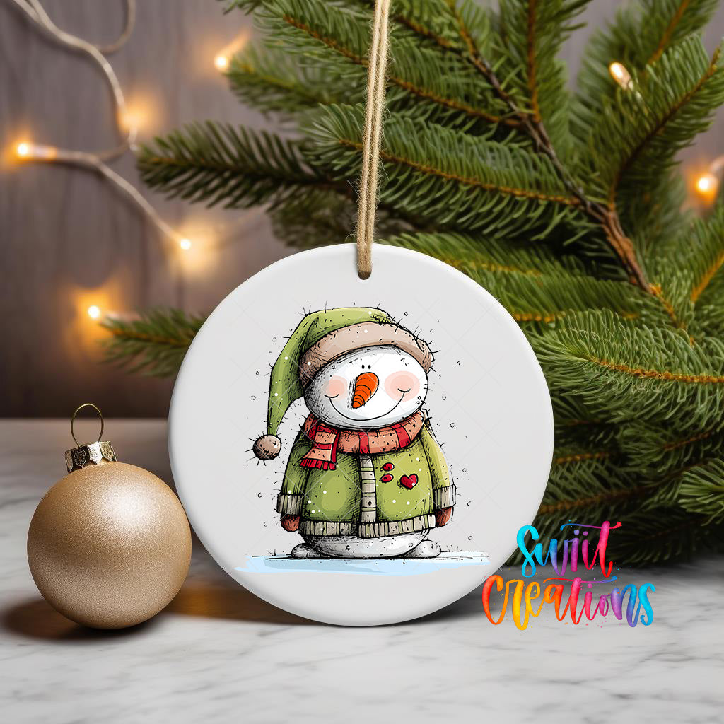 A white ceramic ornament featuring a snowman with a green hat and scarf, sitting on a white surface next to a gold ornament and a Christmas tree with twinkling lights.