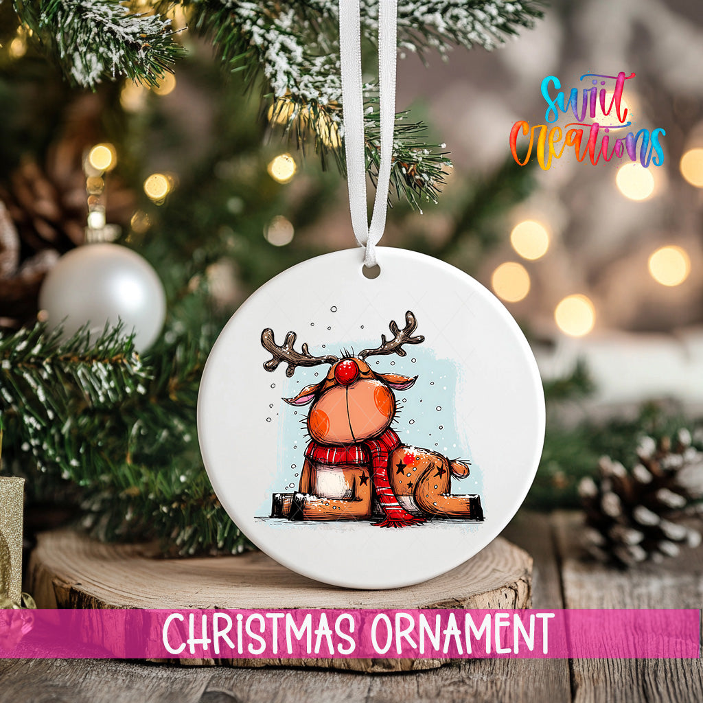A white Christmas ornament featuring a cartoon reindeer is hanging from a ribbon, with a blurred background of a decorated Christmas tree and other ornaments.
