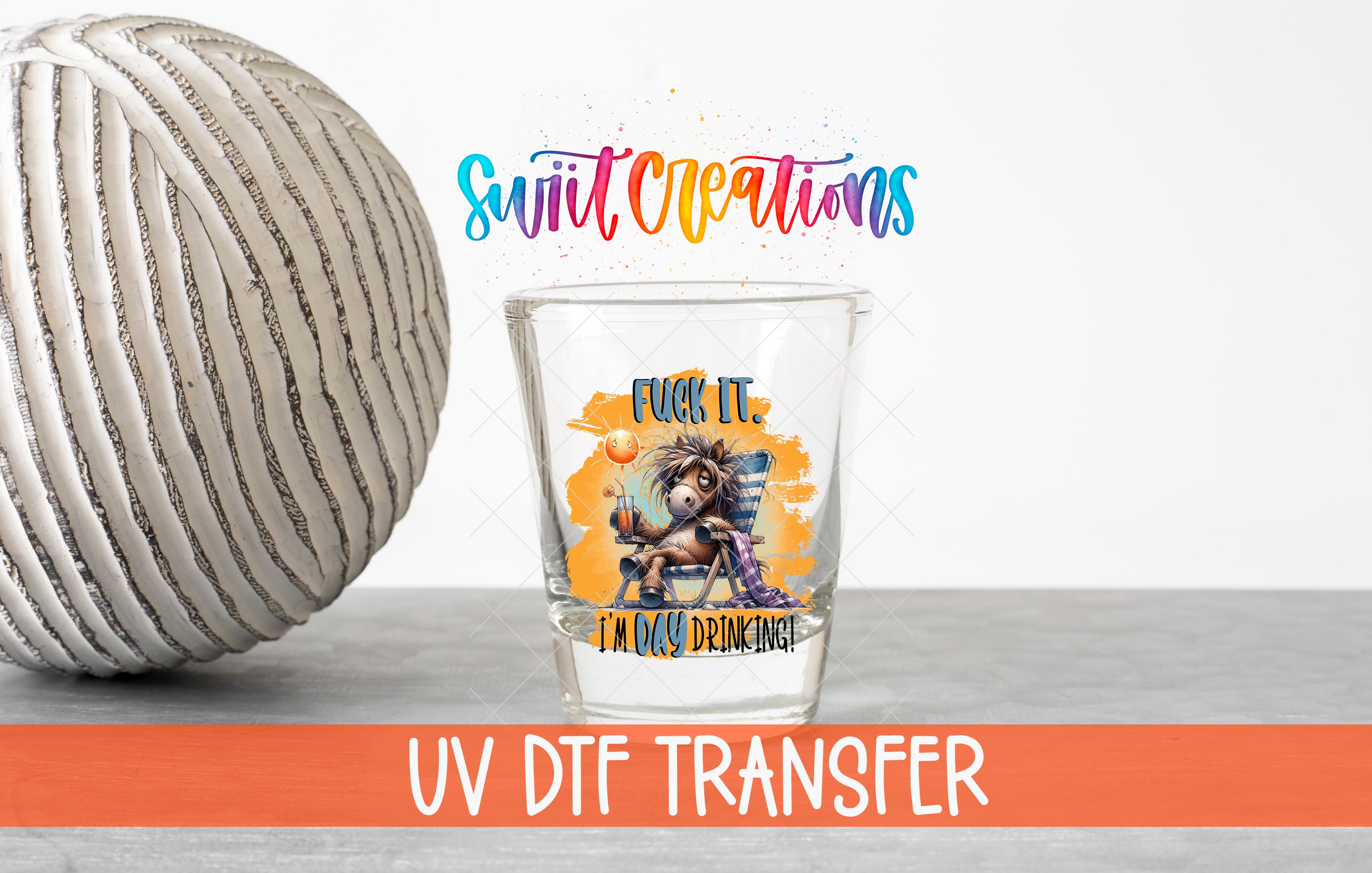 a shot glass with a colorful design and text, placed next to a white ball on a gray surface.