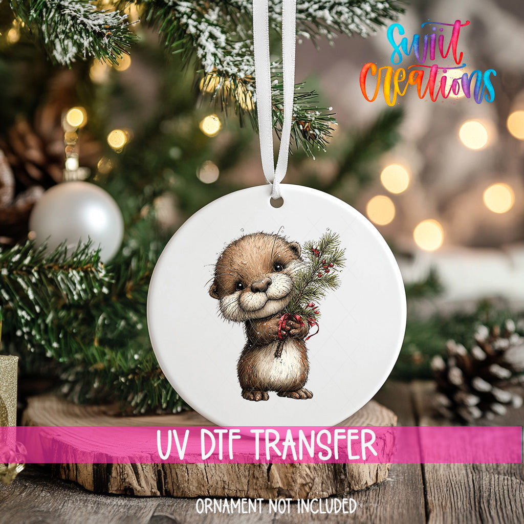 A white ornament with a cute otter illustration is hanging on a Christmas tree, surrounded by pine branches and other holiday decorations.