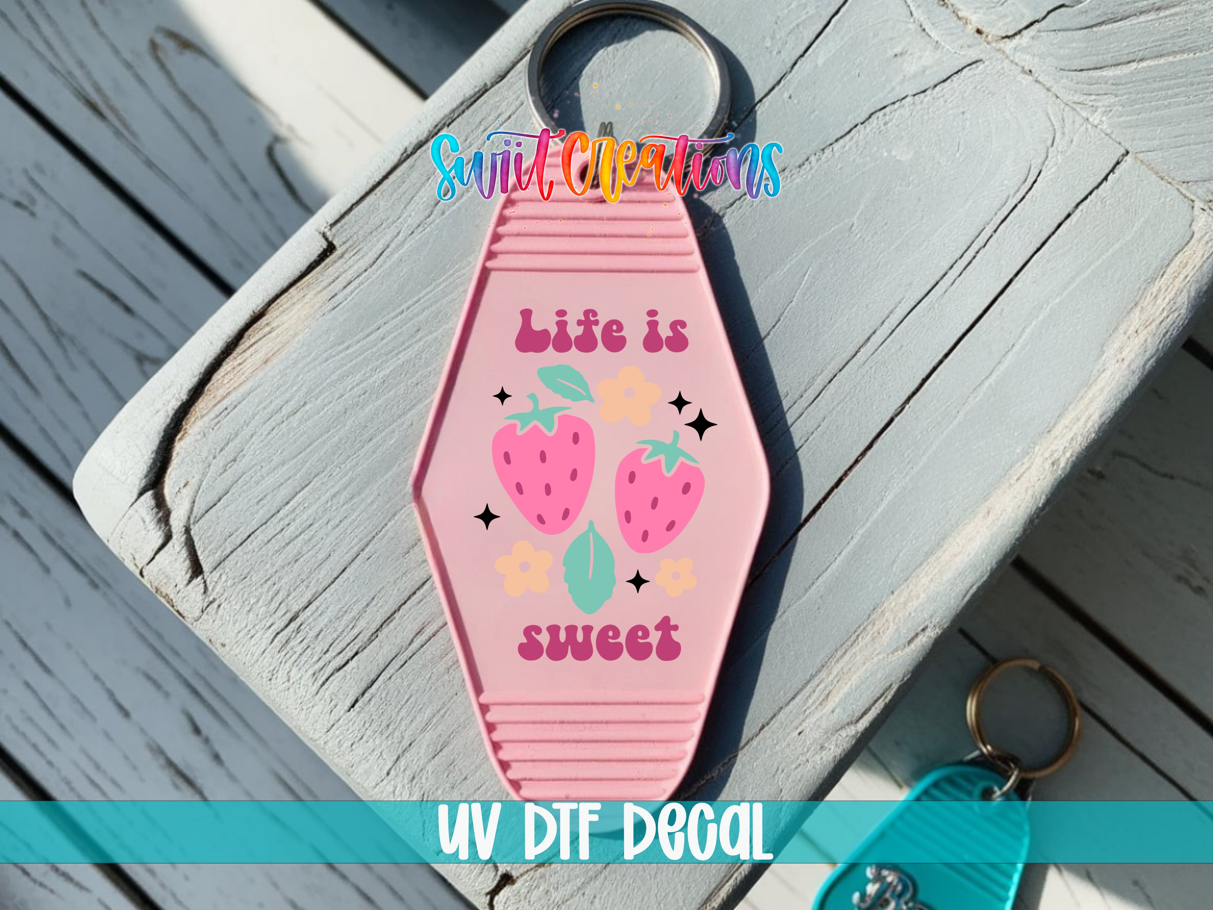 A pink keychain with a design featuring strawberries and the text "Life is sweet" is shown on a wooden surface.