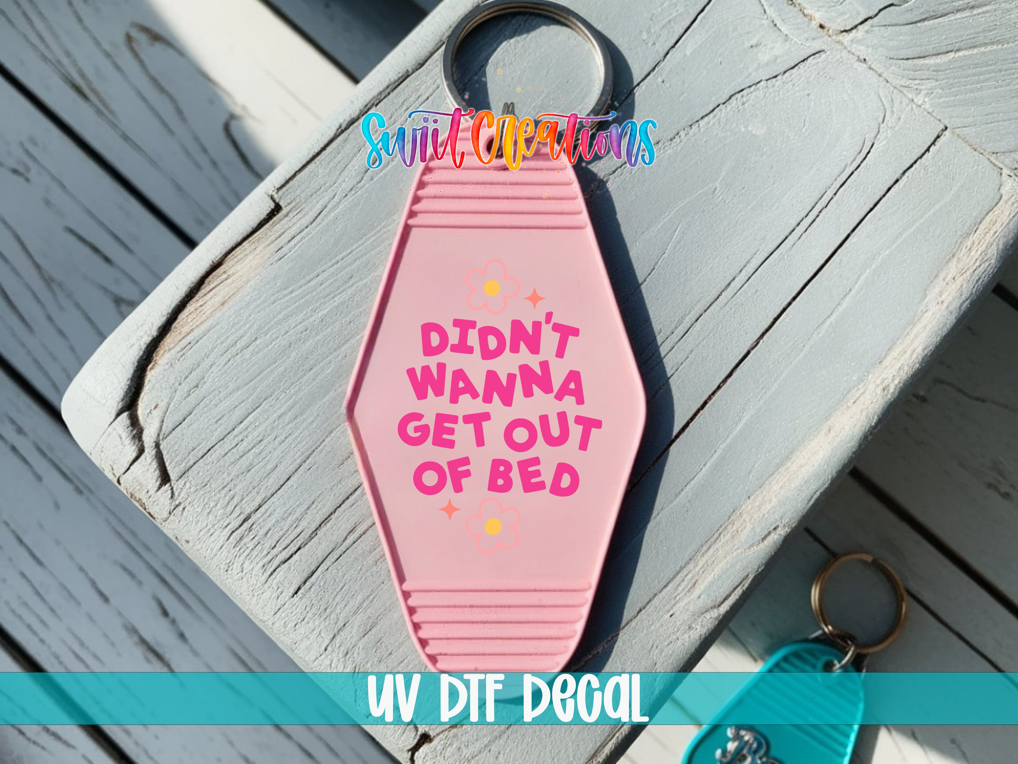 A pink keychain with the text "DIDN'T WAINA GET OUT OF BED" printed on it, placed on a wooden surface.
