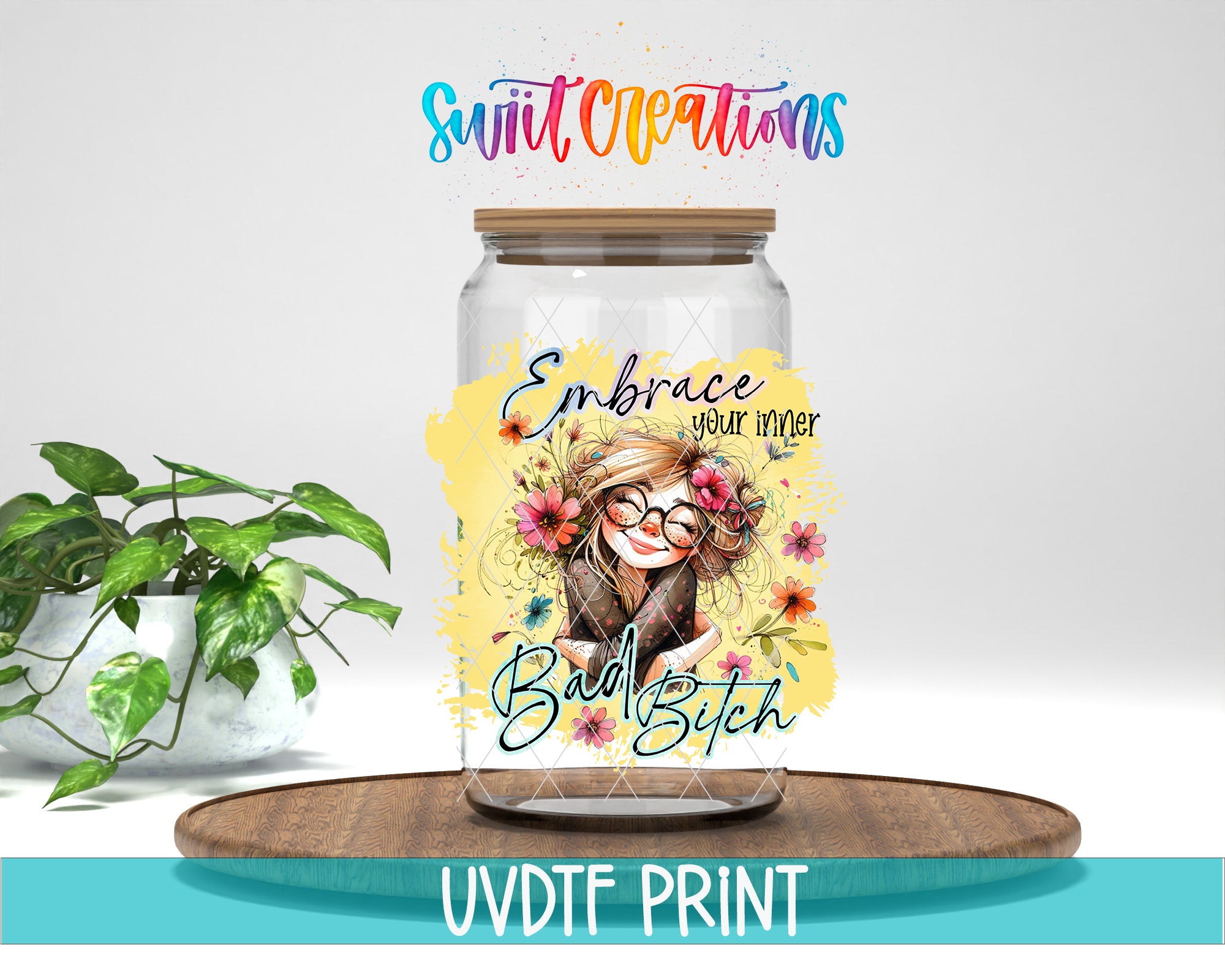 a glass jar with a colorful design, including a woman's face and flowers, along with the text "Embrace your inner Bad Bitch" printed on it. The jar is placed on a wooden surface, and there is a potted plant in the background.