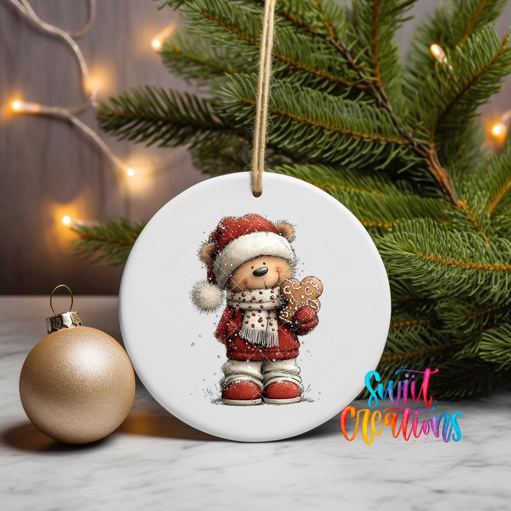 A white ceramic ornament featuring a teddy bear wearing a Santa hat and holding a gingerbread man, placed on a marble surface next to a gold ornament and a Christmas tree.