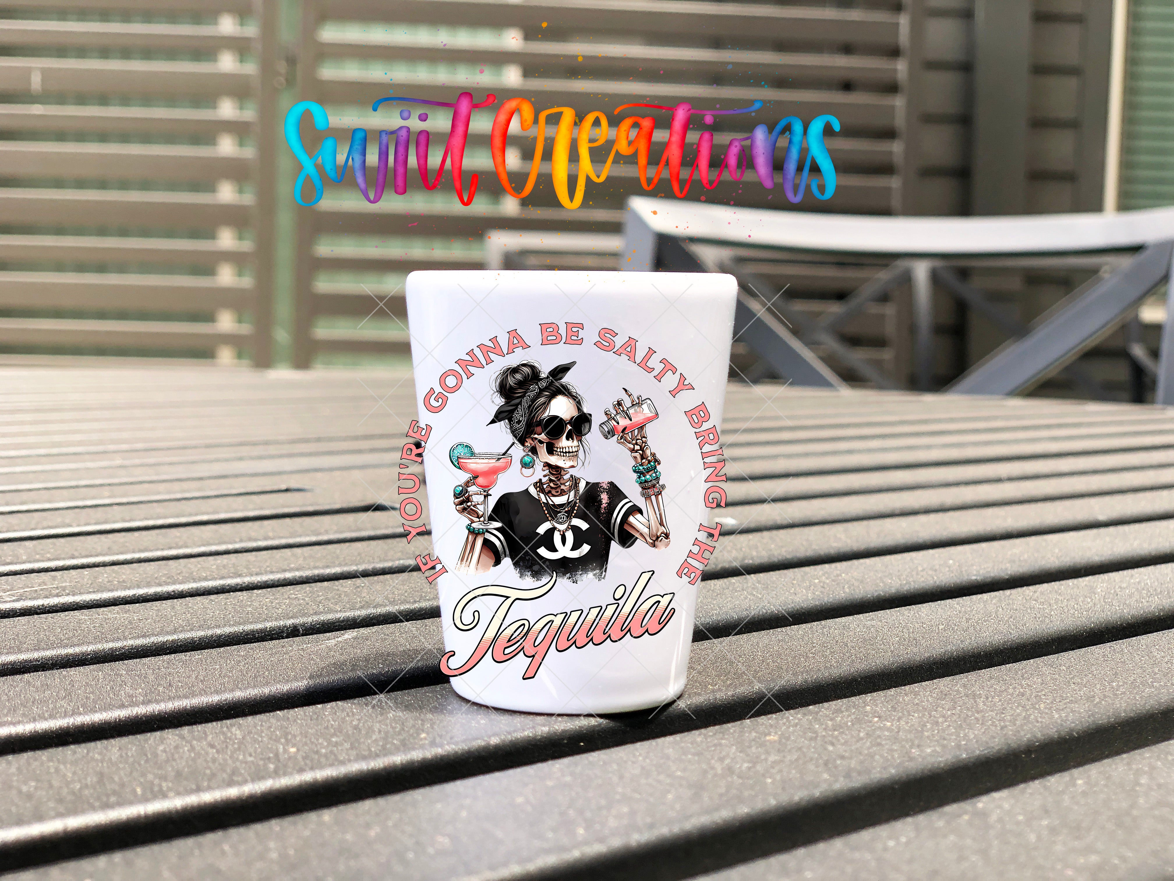 A white shot glass with a colorful design and text on it, placed on a wooden surface.