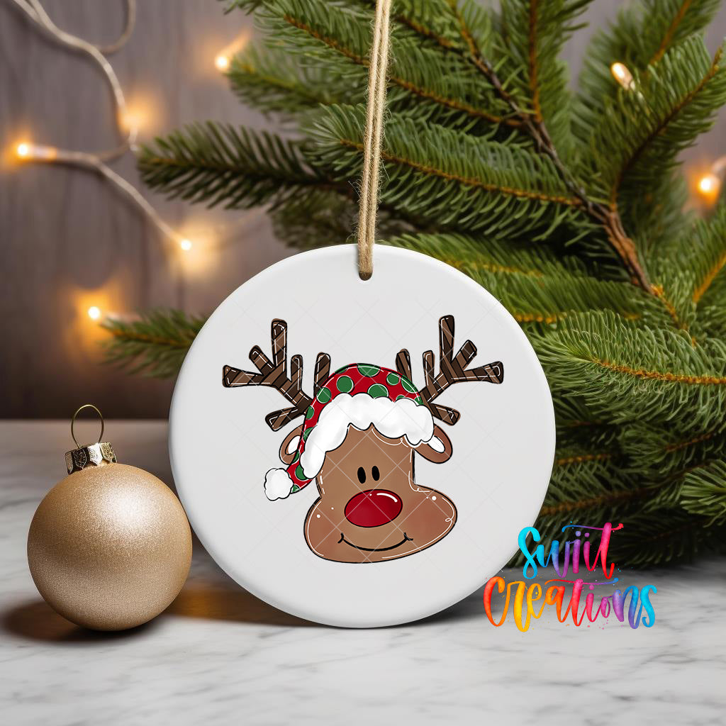 A white ornament with a cartoon reindeer wearing a Santa hat is displayed on a table, with a Christmas tree and twinkling lights in the background.