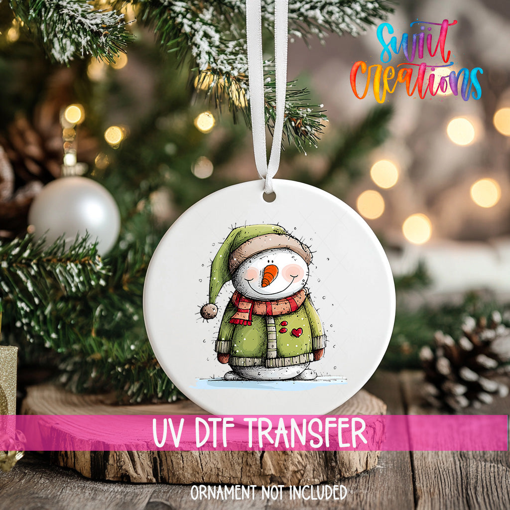 A white ornament with a snowman design hangs on a Christmas tree, surrounded by pine branches and twinkling lights.