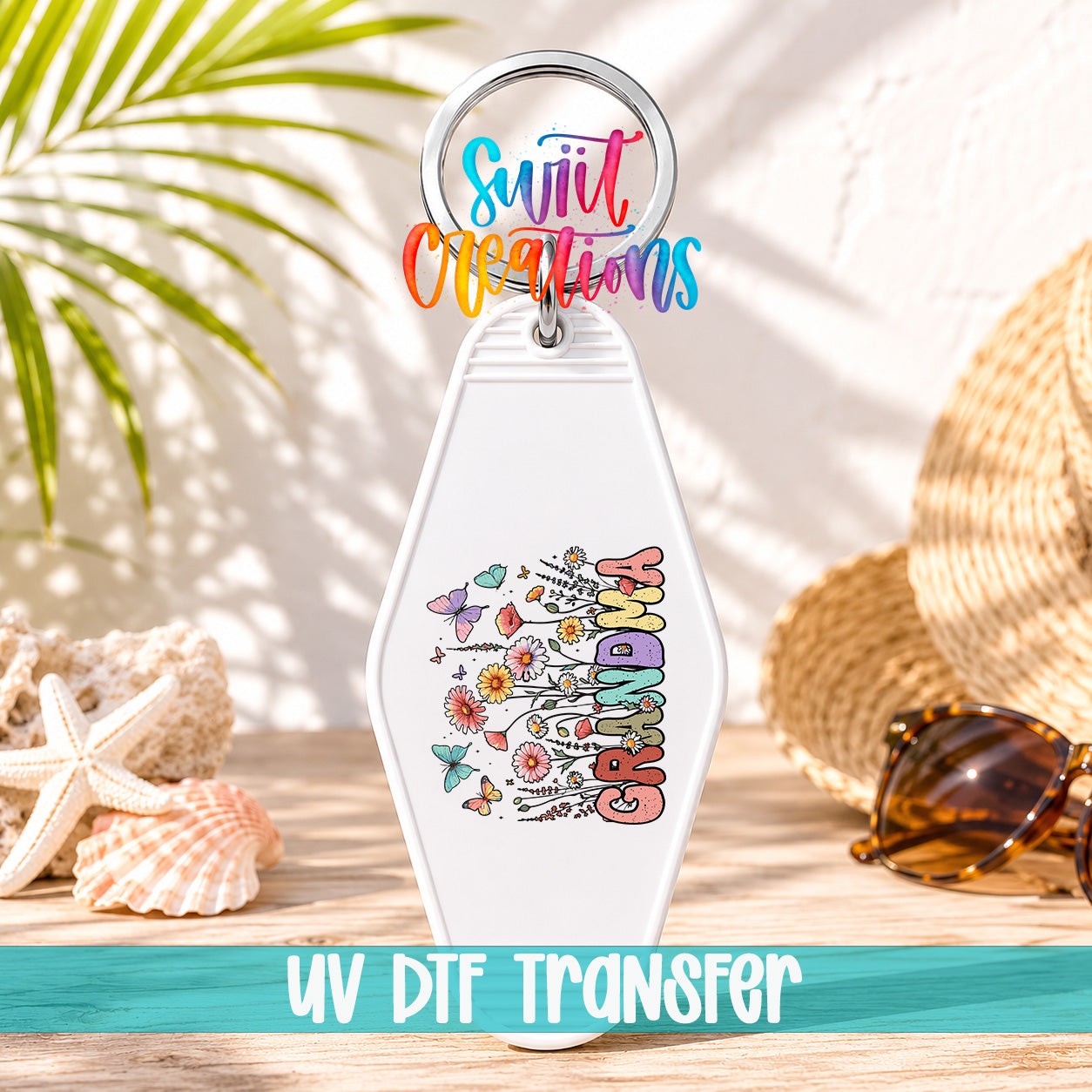 A keychain with a colorful design featuring the word "Grandma" surrounded by various flowers and butterflies. The keychain is placed on a wooden surface, with sunglasses and a straw hat visible in the background.