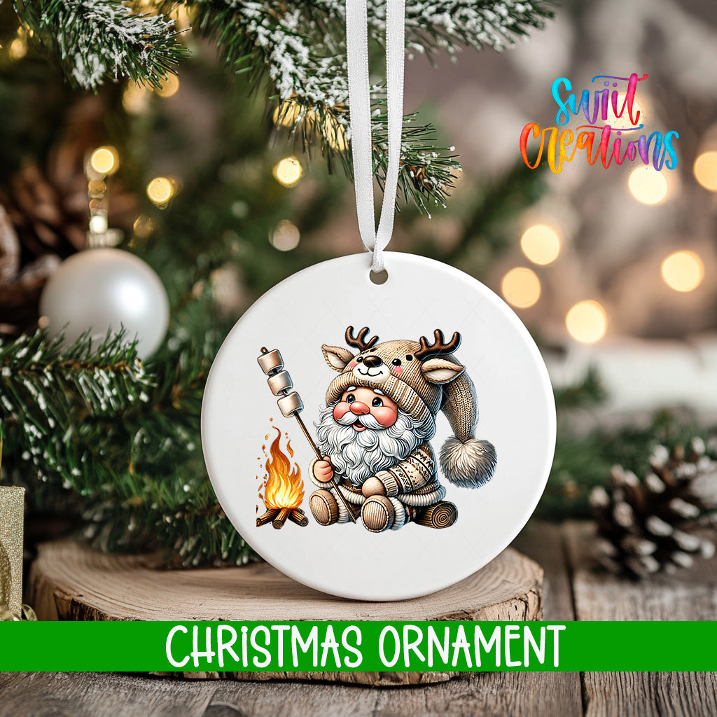 A white Christmas ornament featuring a cartoon Santa Claus holding a marshmallow over a campfire, with a pine tree and twinkling lights in the background.