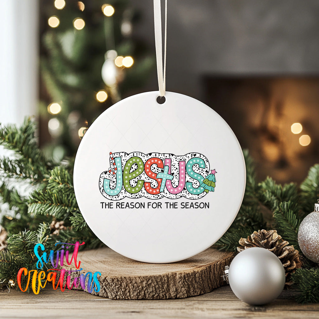 A white ceramic ornament with the word "JESUS" and the phrase "THE REASON FOR THE SEASON" printed on it, hanging on a string. The ornament is surrounded by Christmas decorations, including ornaments and pine cones, on a wooden surface.