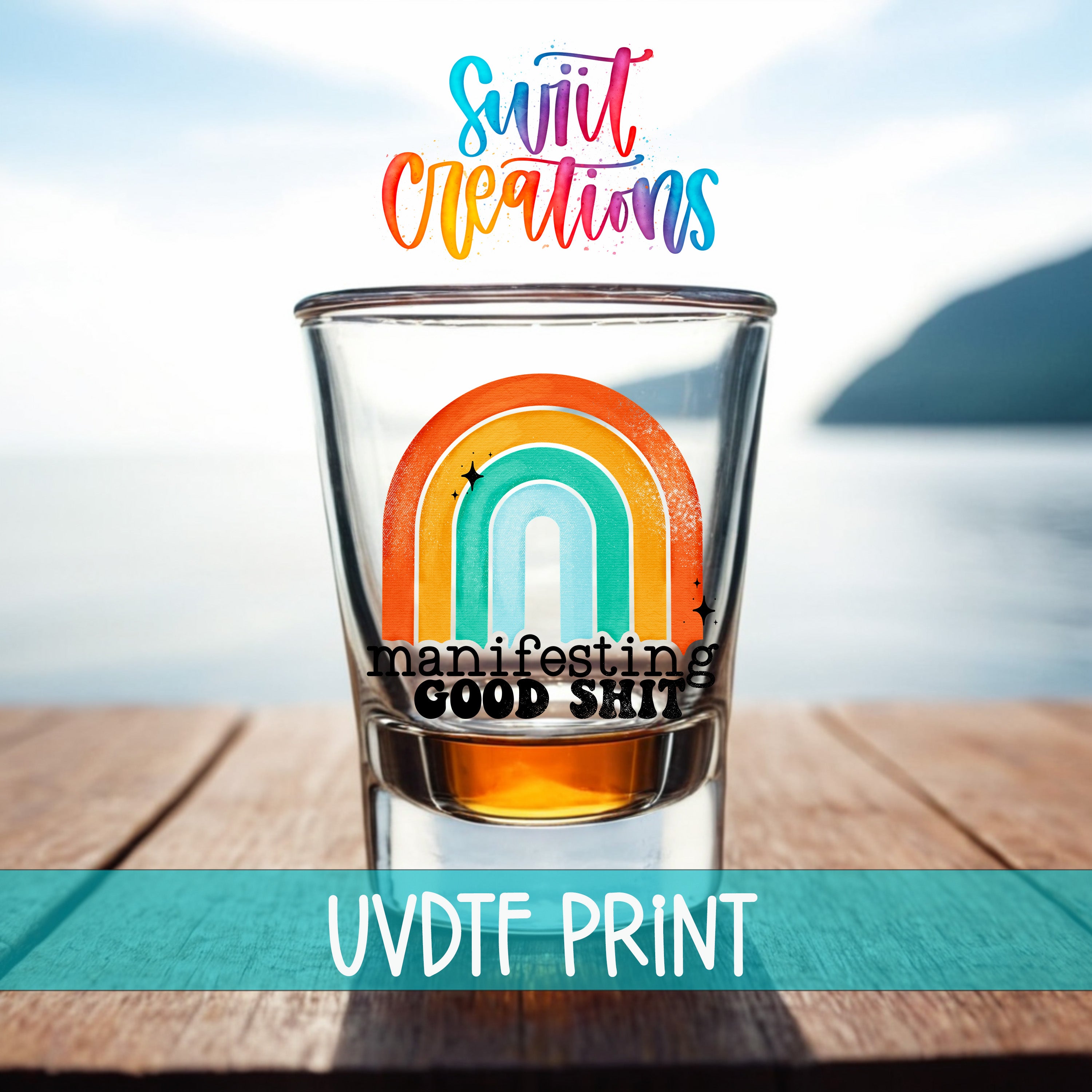 A shot glass with a rainbow graphic and the text "Manifesting GOOD SHIT" printed on it, placed on a wooden surface with a body of water in the background.
