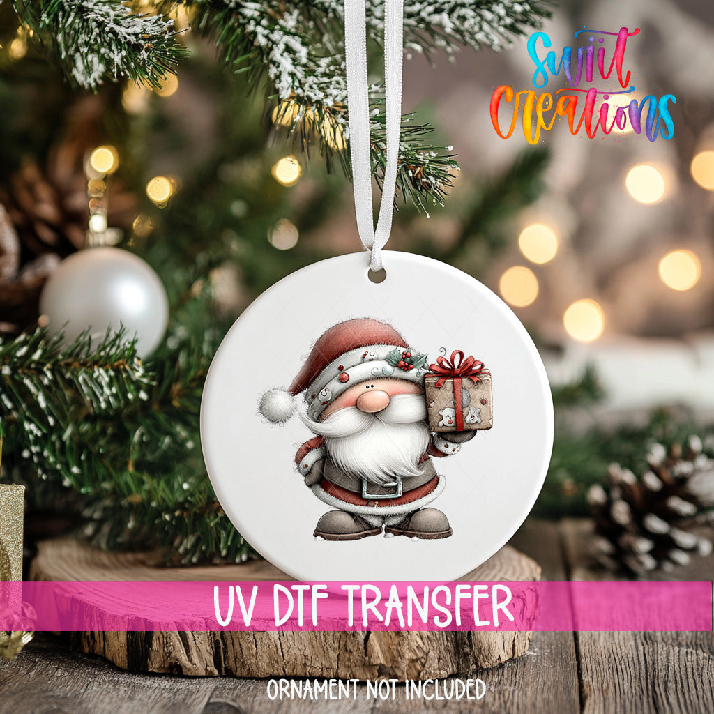 A white ornament with a Santa Claus figure holding a gift is hanging on a Christmas tree.