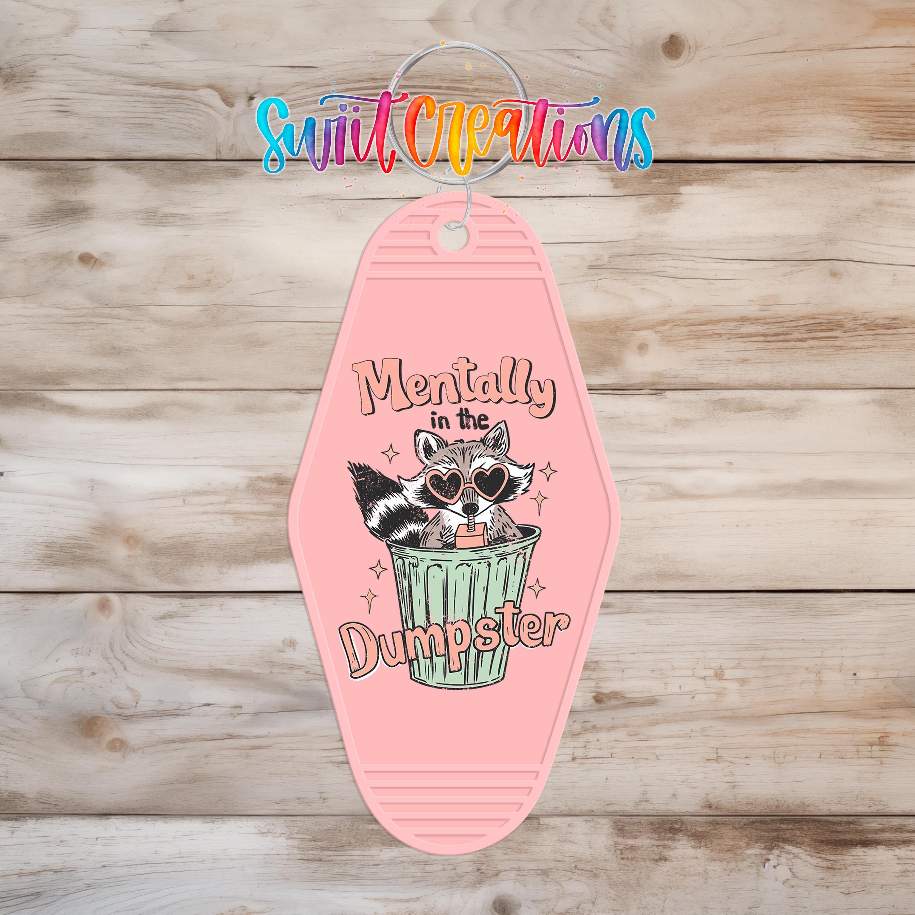 A pink keychain with a cartoon raccoon and the text "Mentally in the Dumpster" printed on it.
