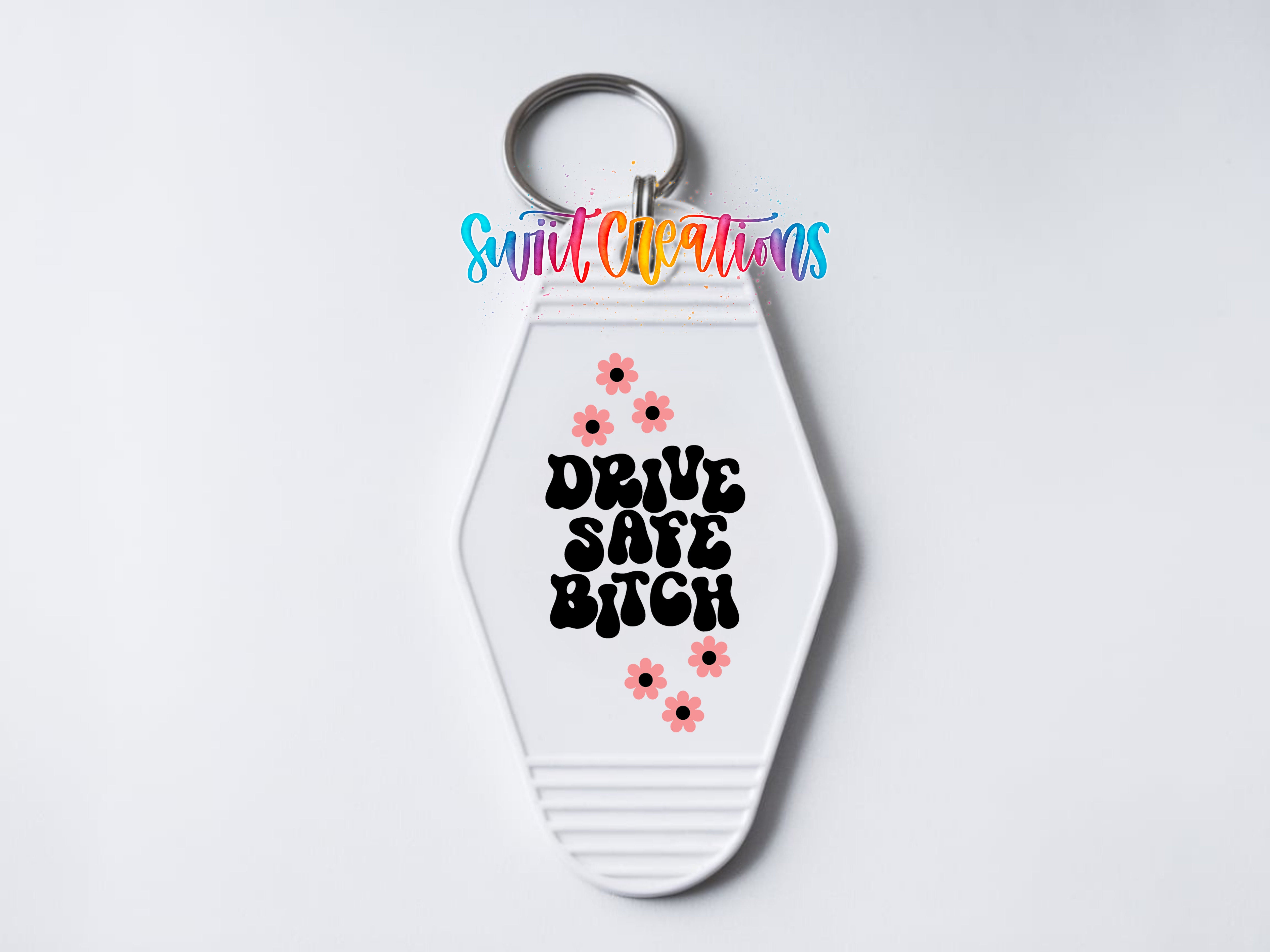 A keychain with a white hexagonal shape and a keyring attached to it. The keychain has a message printed on it in a stylized font, which reads "DRIVE SAFE BITCH" along with some pink flowers.