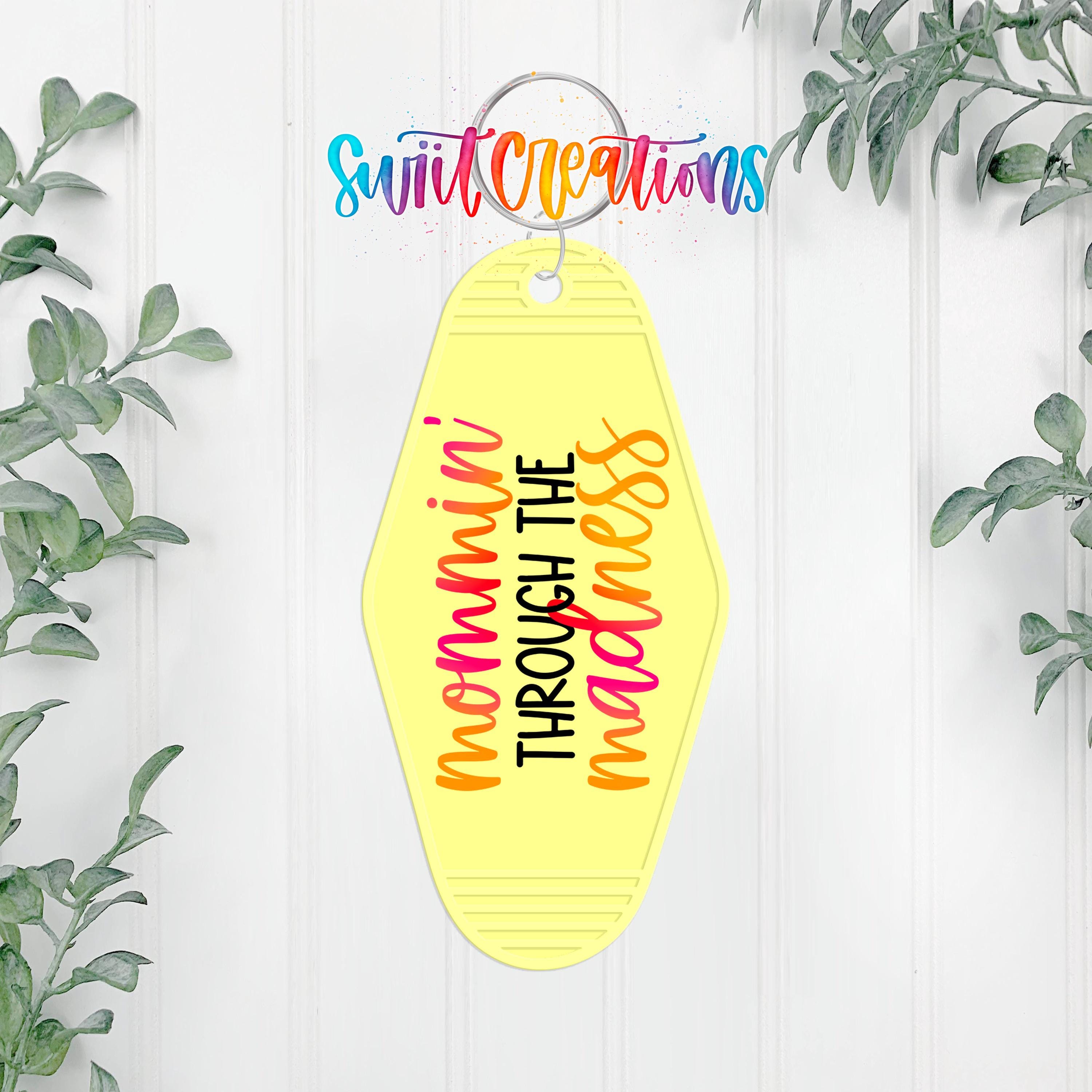 A yellow keychain with the text "momma through the mess" printed on it, hanging on a white wall with greenery in the background.