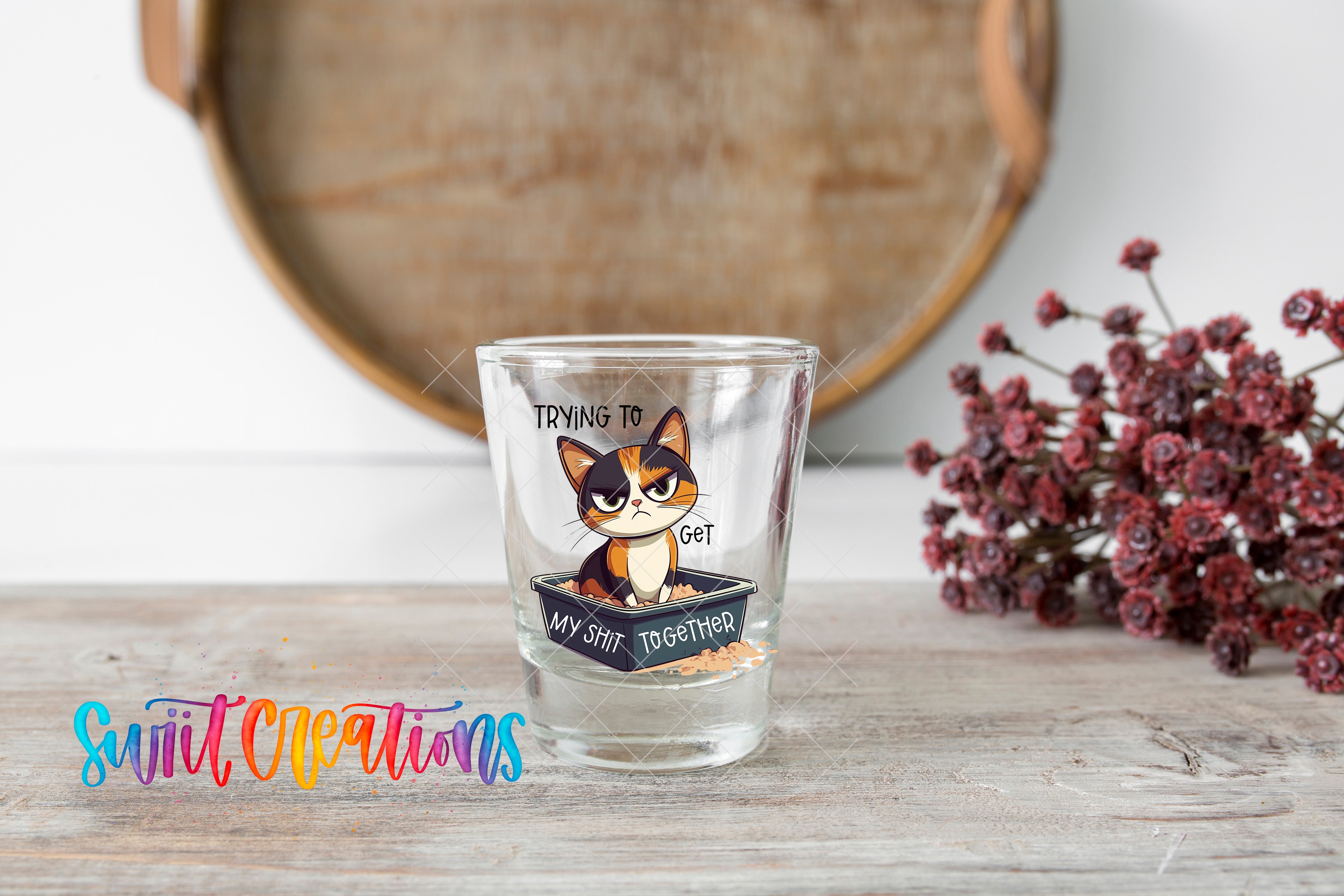 A shot glass with a cat illustration and the text "Trying to get my cat together" is placed on a wooden surface, with a bouquet of flowers and a wooden tray in the background.