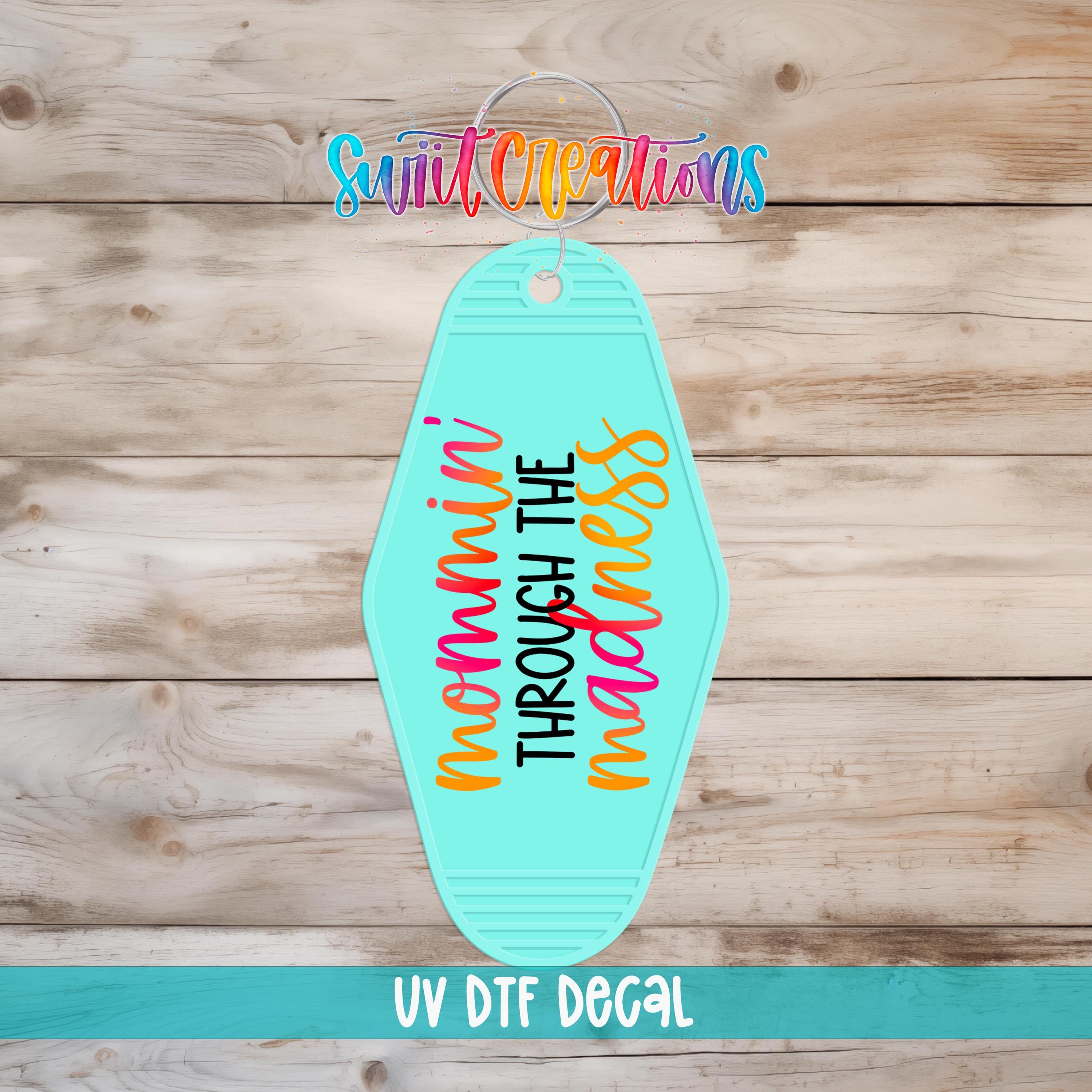 A turquoise keychain with the text "MOMA HIN'T THROUGH THE MAGIC" printed on it, along with a colorful logo and the text "UV Dtf Decal" at the bottom.