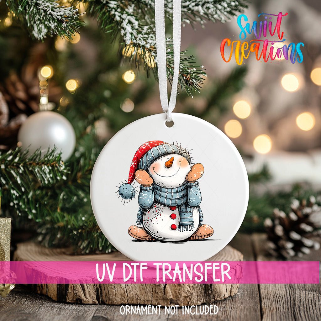 A white ornament with a snowman design hangs on a Christmas tree, surrounded by pine branches and twinkling lights.