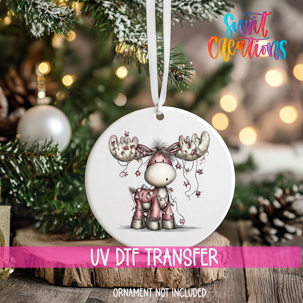 A white ornament with a cartoon moose and Christmas lights is hanging on a tree, with a pink ribbon around it.