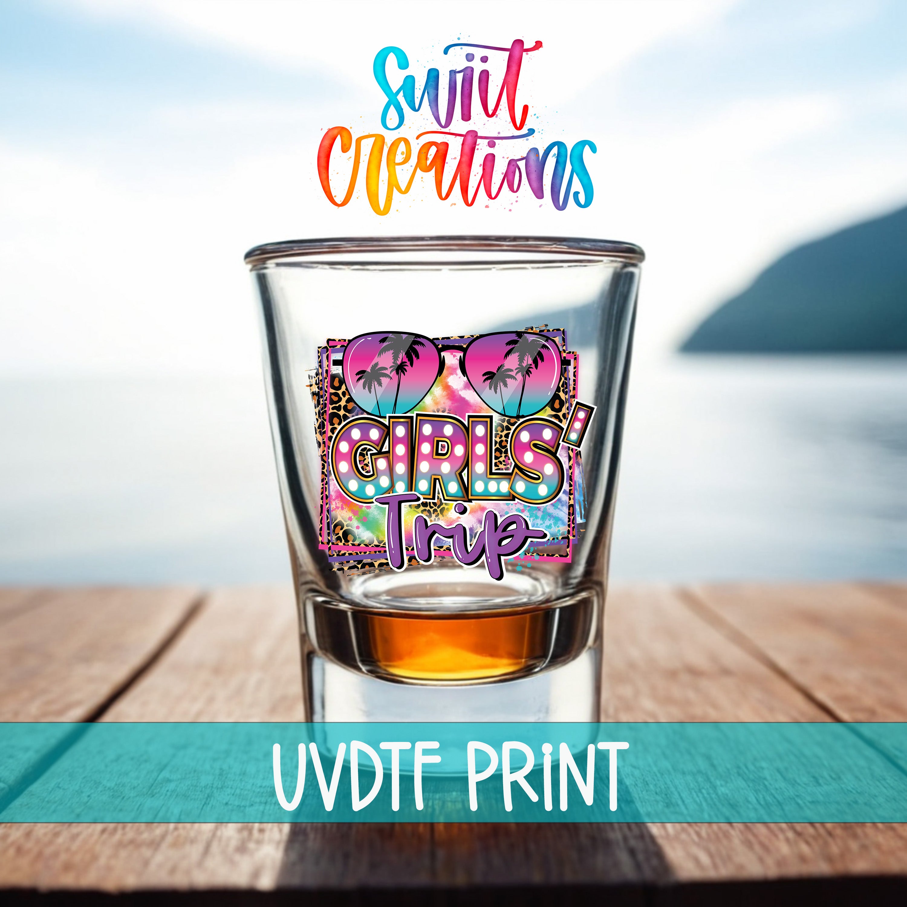 A colorful cocktail glass with a graphic design featuring sunglasses and the text "Girls Trip" printed on it, placed on a wooden surface with a body of water in the background.