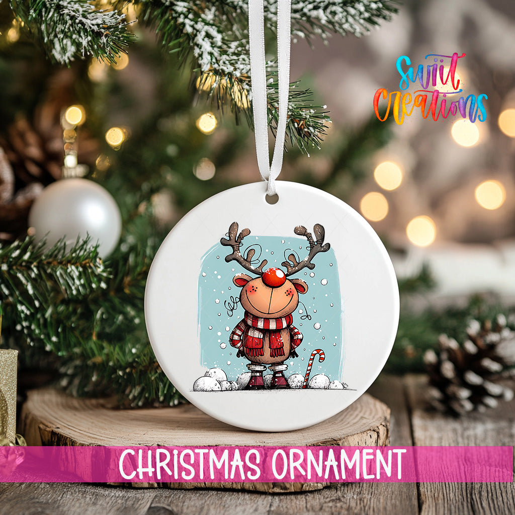 A white Christmas ornament featuring a cartoon reindeer with antlers and a red nose, hanging on a tree branch with snowflakes falling around it.