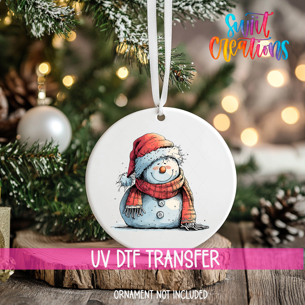 A white ornament with a snowman design hangs on a Christmas tree, surrounded by pine branches and twinkling lights.