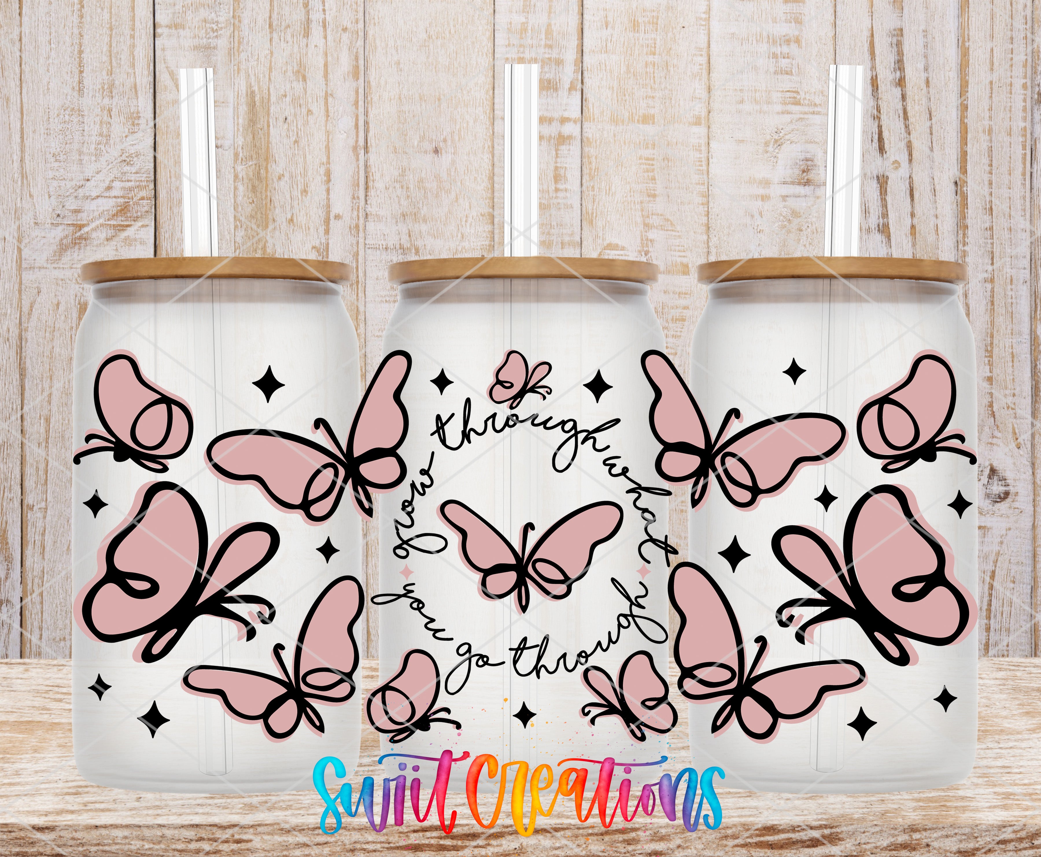 Three glass tumblers with a butterfly design, each featuring a straw. The tumblers are arranged in a row, with the middle one slightly larger than the other two. The background is a wooden surface with a textured appearance.