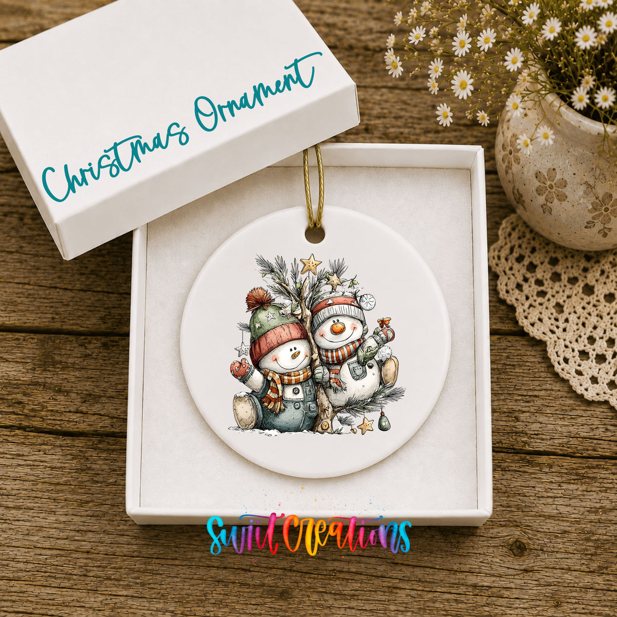 A white Christmas ornament with a snowman and pine tree design is displayed in a white box, accompanied by a potted plant and a lace doily on a wooden surface.