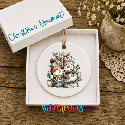 A white Christmas ornament with a snowman and pine tree design is displayed in a white box, accompanied by a potted plant and a lace doily on a wooden surface.
