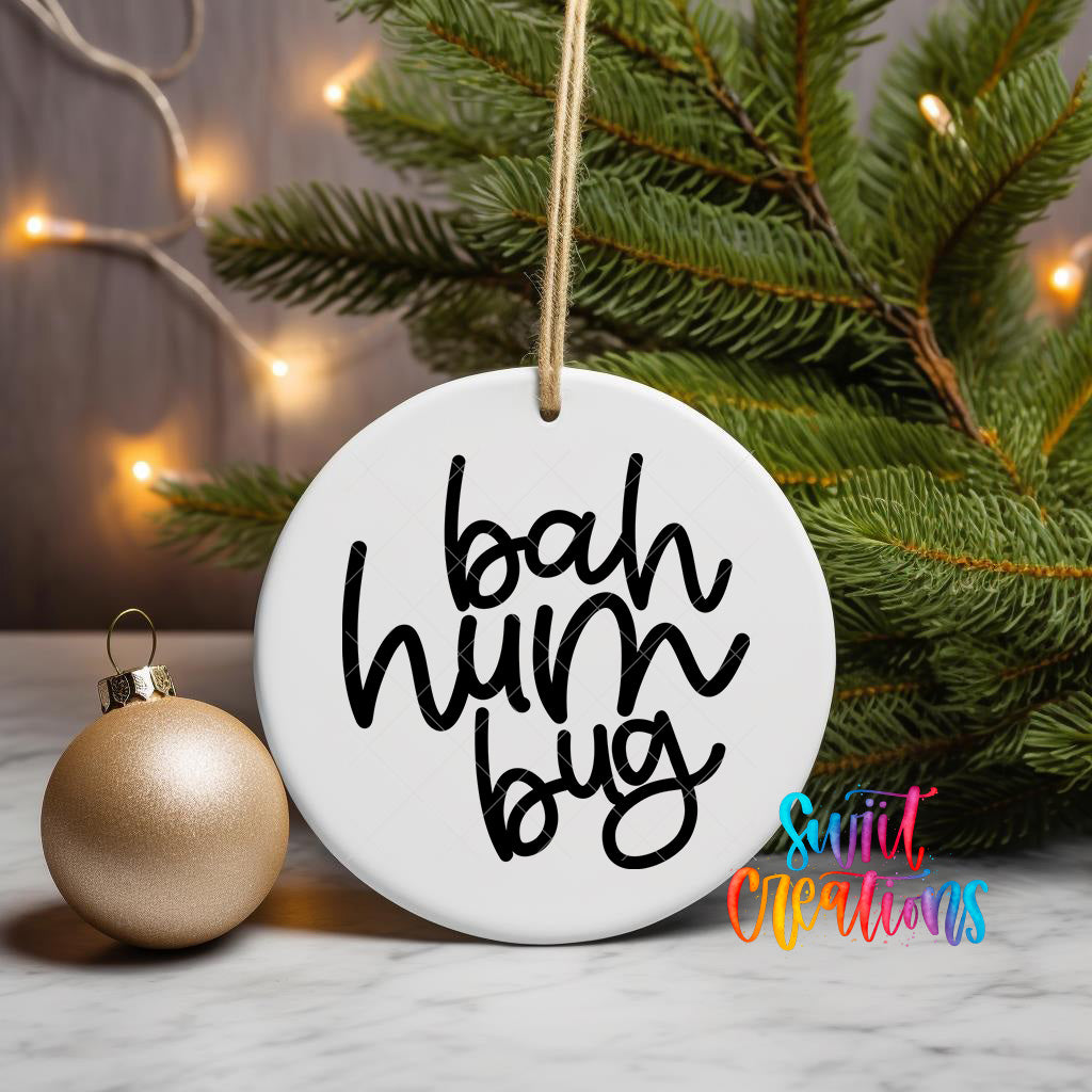 A white ornament with the text "bah hum bug" written on it, hanging from a string, is placed next to a gold ornament and a pine tree with twinkling lights in the background.