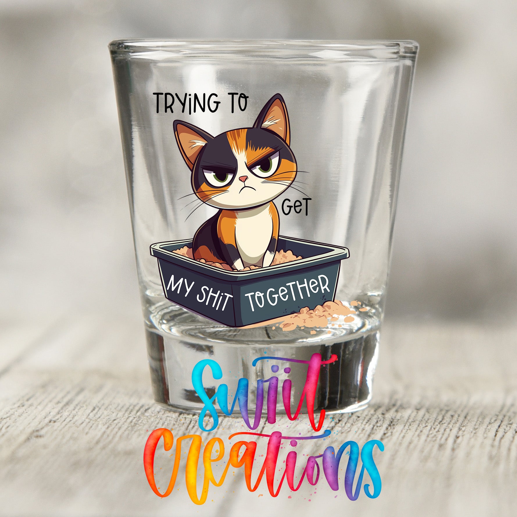 A shot glass with a cartoon cat sitting in a litter box and the text "TRYING TO GET MY SHIT TOGETHER" printed on it.