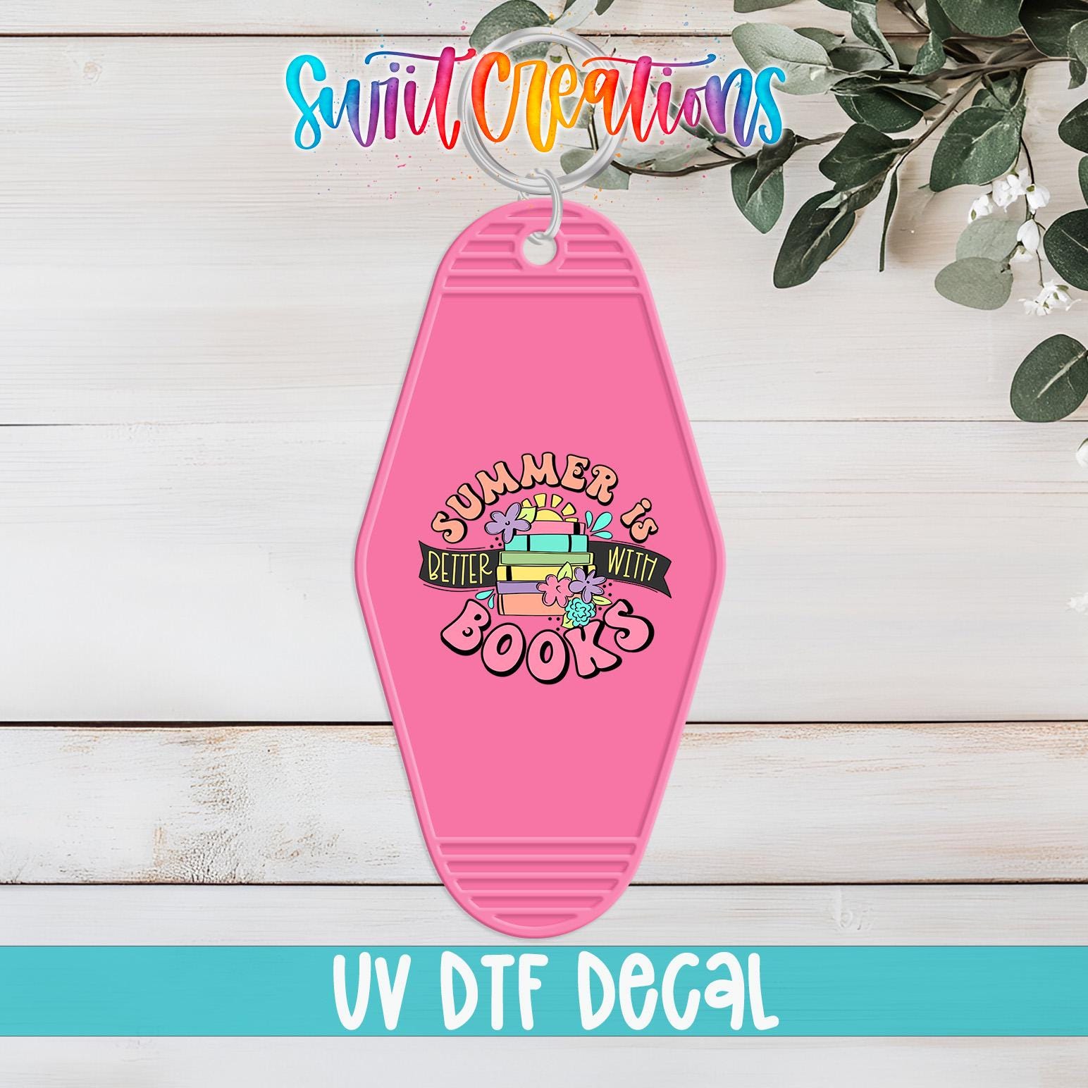 A pink keychain with a colorful design and text, placed on a wooden surface with greenery in the background.