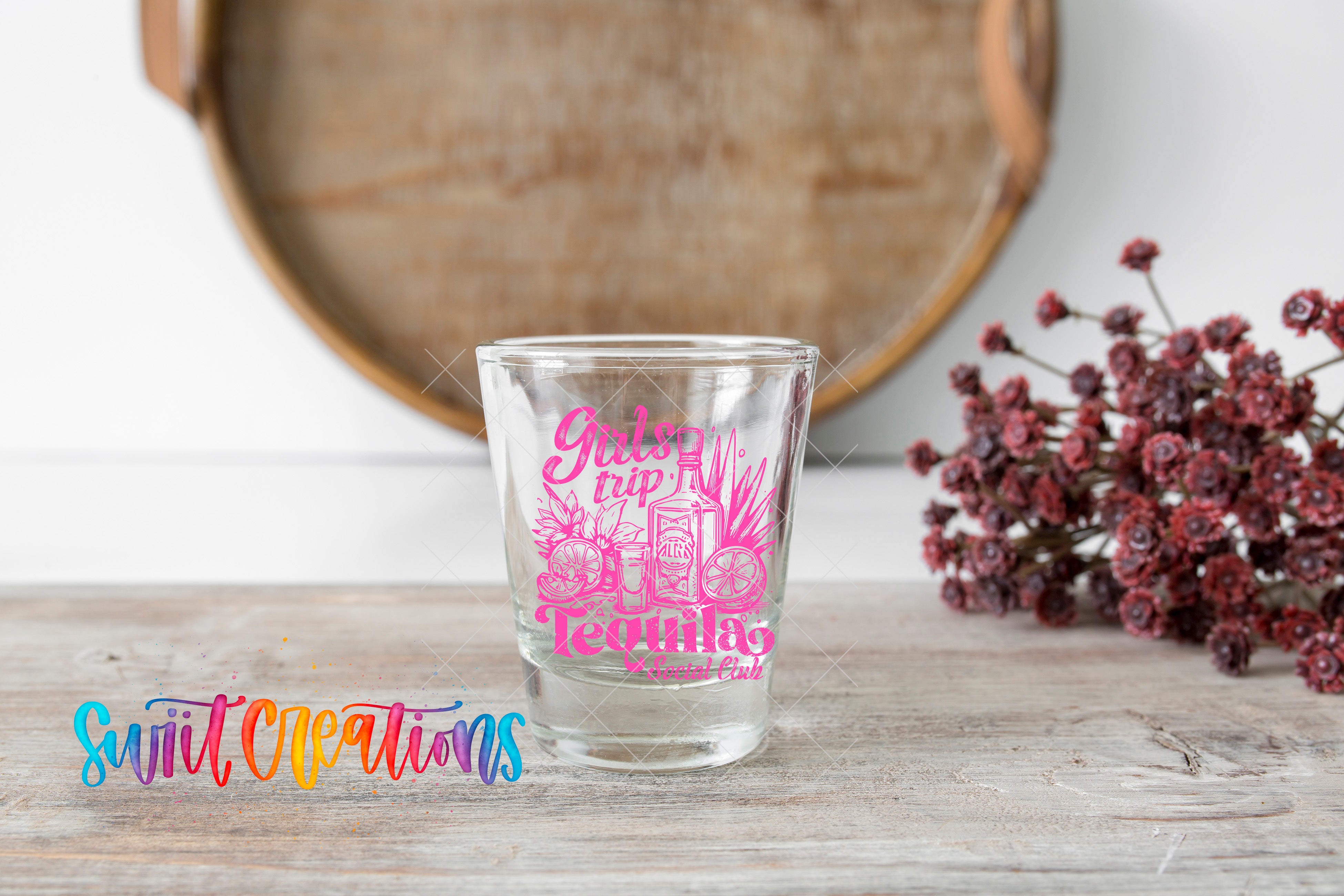 A shot glass with a pink design and text is placed on a wooden surface, with a bunch of red flowers and a wooden tray in the background.