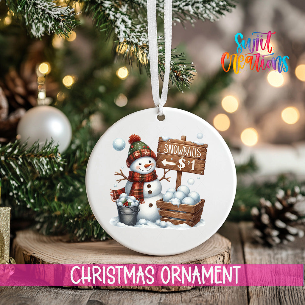 A white Christmas ornament featuring a snowman holding a sign that says "Snowballs $1" is hanging on a tree branch.