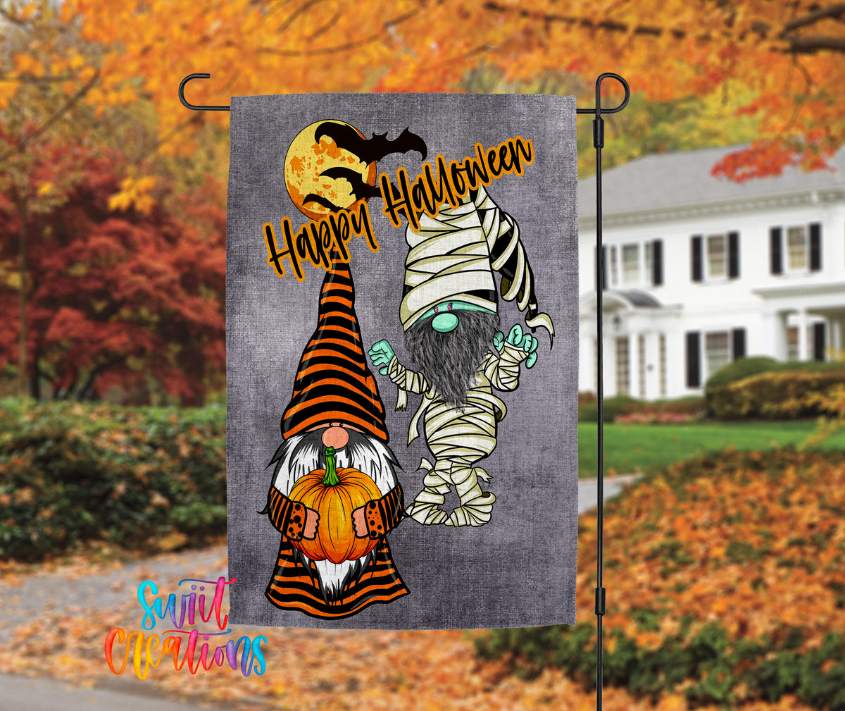 A Halloween-themed garden flag featuring two gnomes, one dressed as a mummy and the other as a gnome, holding pumpkins and a skeleton. The background is a colorful autumn landscape with a house and trees.