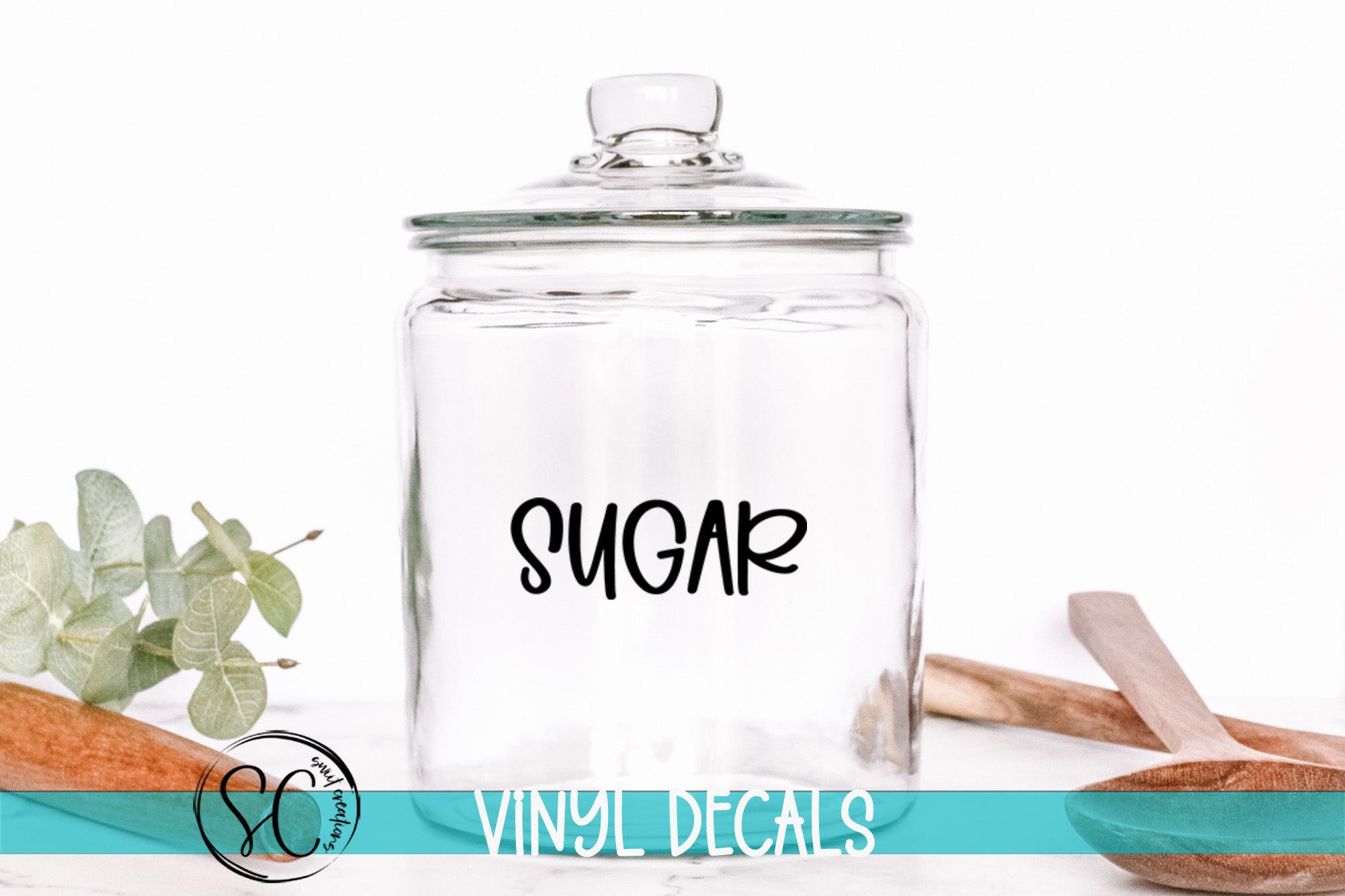 A clear glass jar with the word "SUGAR" printed on it, placed on a white surface next to some wooden utensils.
