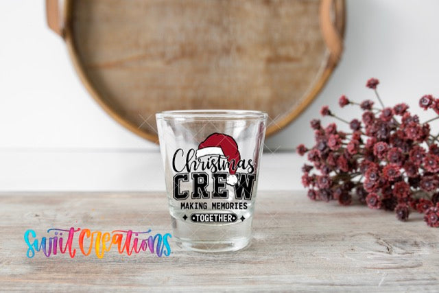 A shot glass with a Christmas crew design sits on a wooden surface, with a bouquet of red flowers and a wooden tray in the background.
