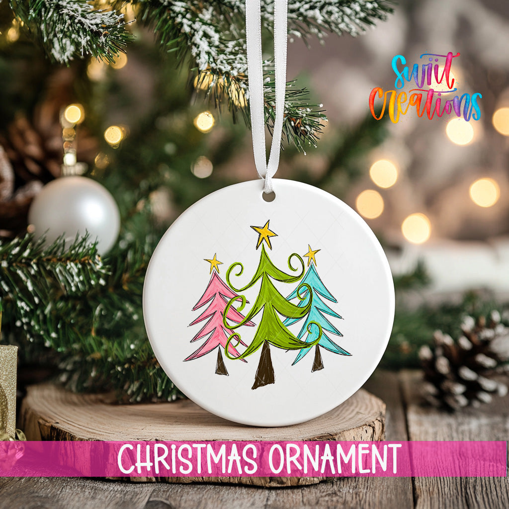 A white Christmas ornament with a colorful illustration of three trees hangs on a tree branch, surrounded by other Christmas decorations and lights.