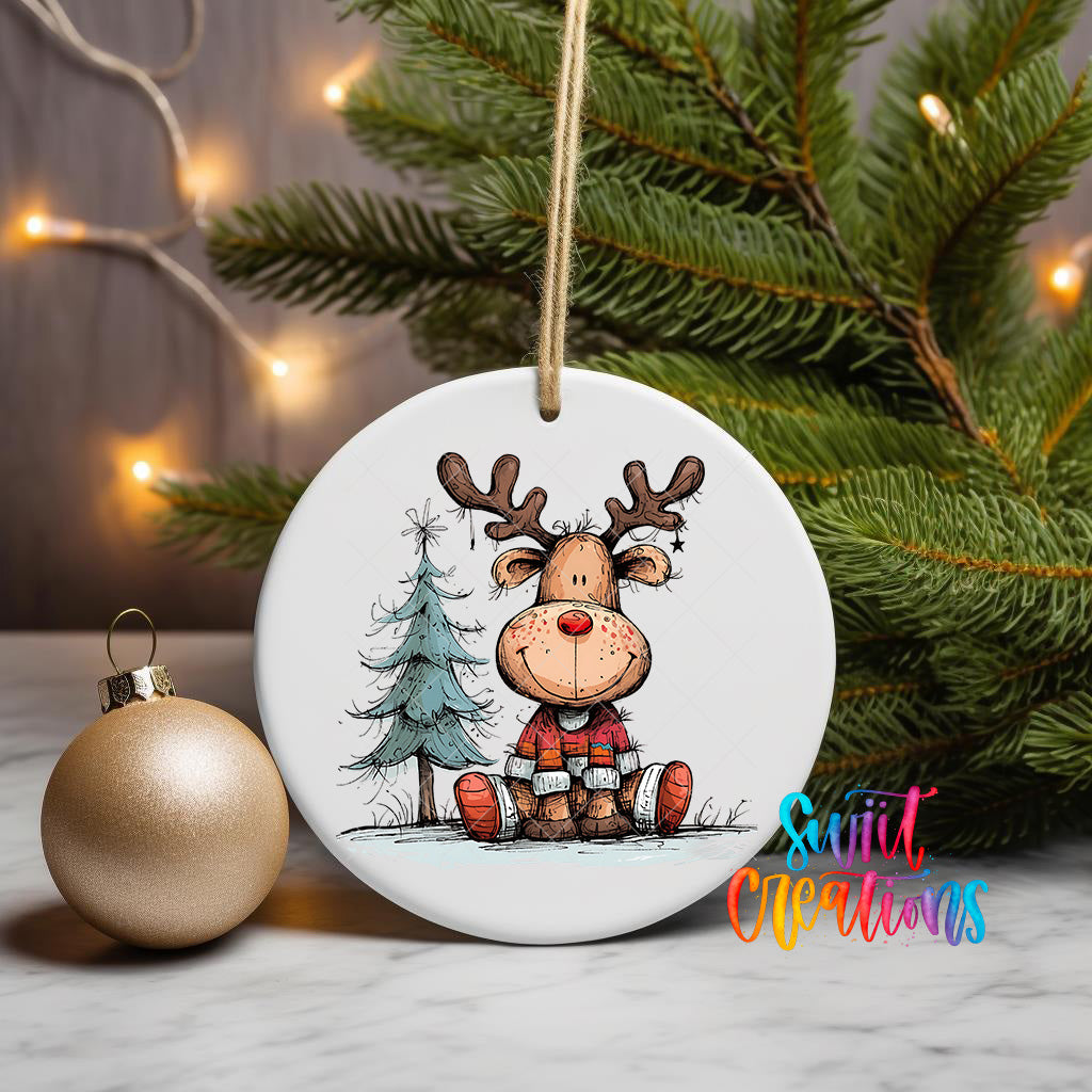 A white ceramic ornament featuring a cartoon reindeer sitting on a snowy landscape, with a Christmas tree and twinkling lights in the background.