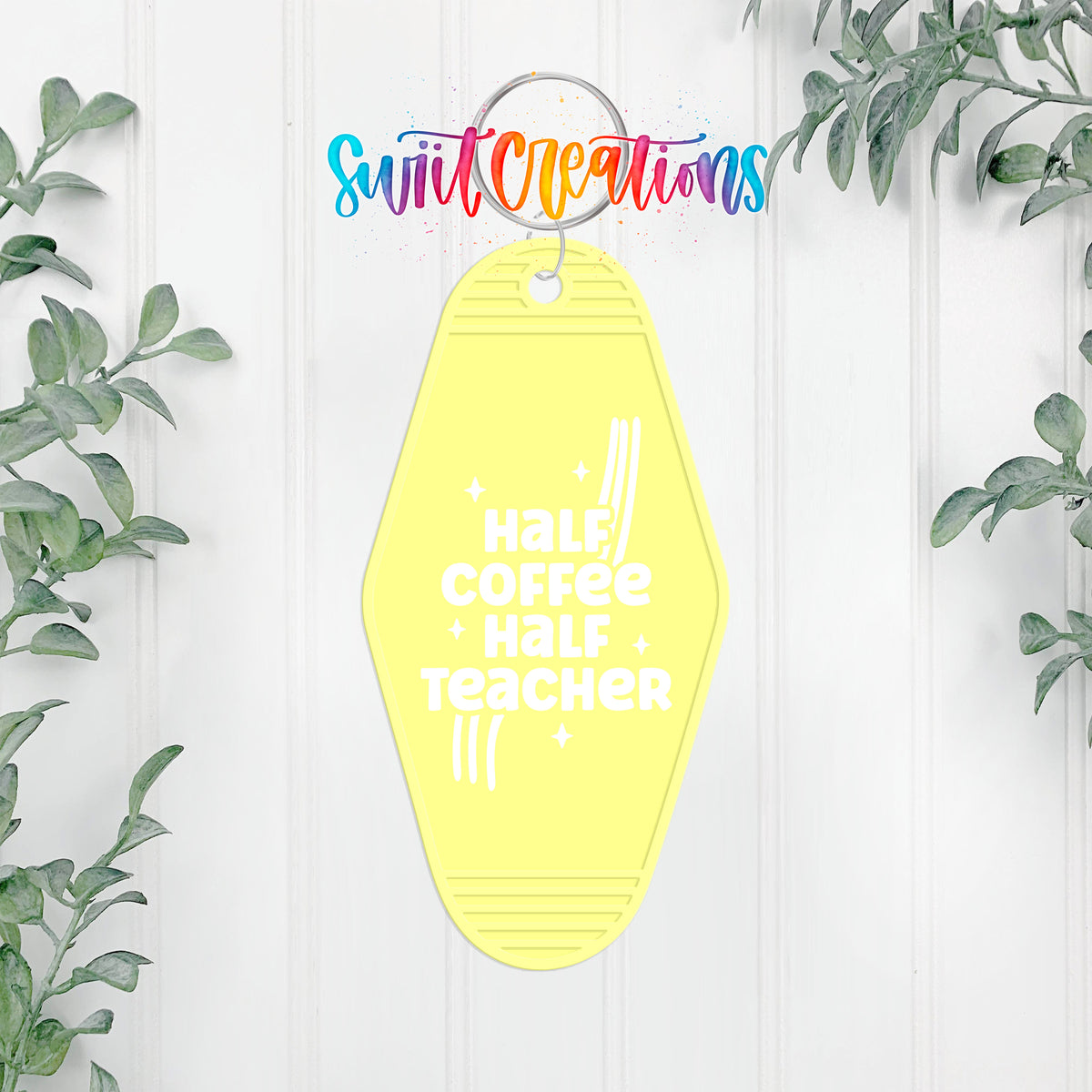 A yellow keychain with the text "Half Coffee Half Teacher" printed on it, hanging on a white wall with greenery in the background.