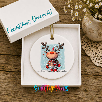 A white Christmas ornament with a reindeer design is displayed in a white box, accompanied by a small potted plant and a lace doily on a wooden surface.