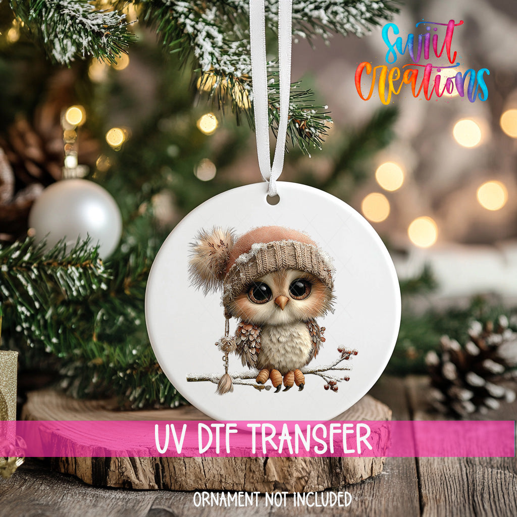 A white ornament with a cute owl wearing a hat and sitting on a branch, hanging on a Christmas tree.