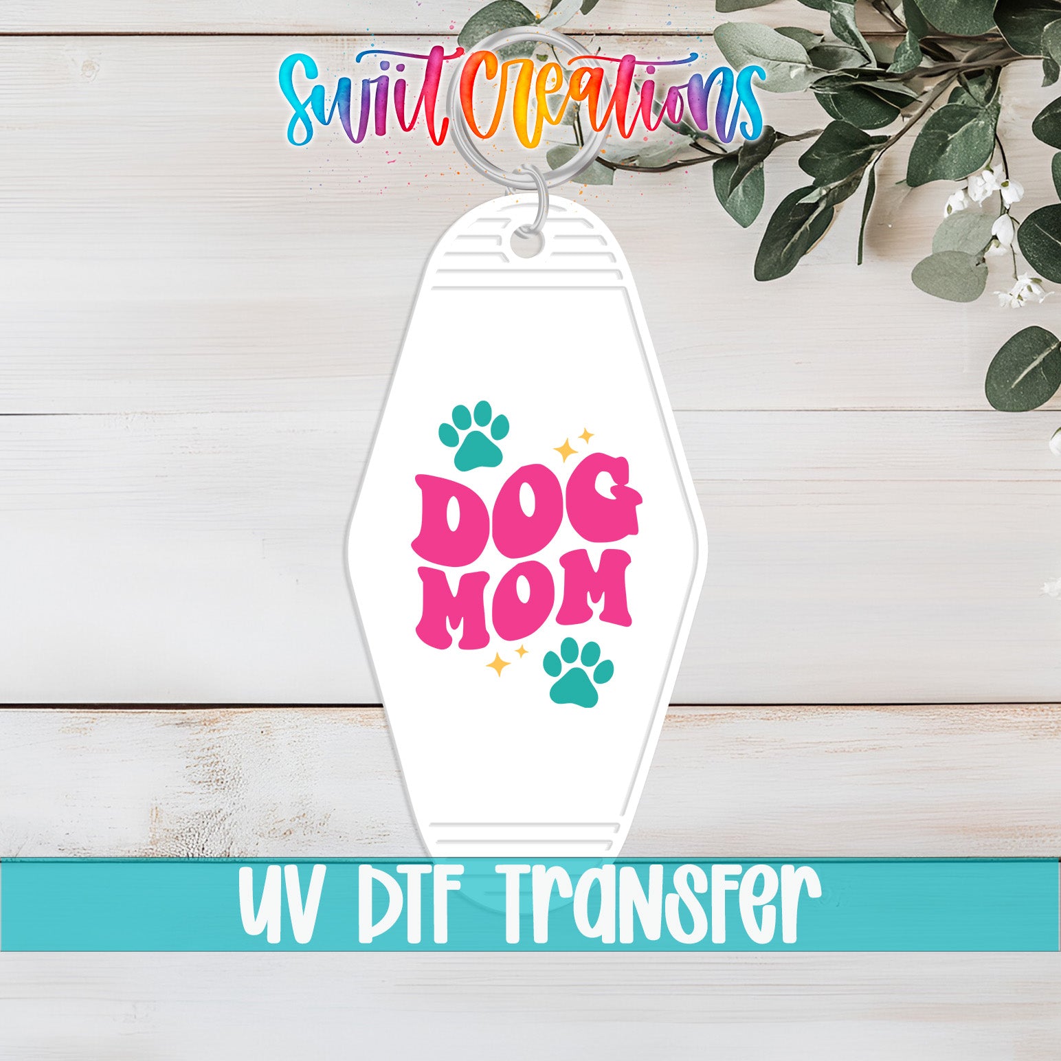 A keychain with the text "DOG MOM" printed on it, along with paw prints and a small star. The keychain is placed on a wooden surface, and there is a plant in the background.