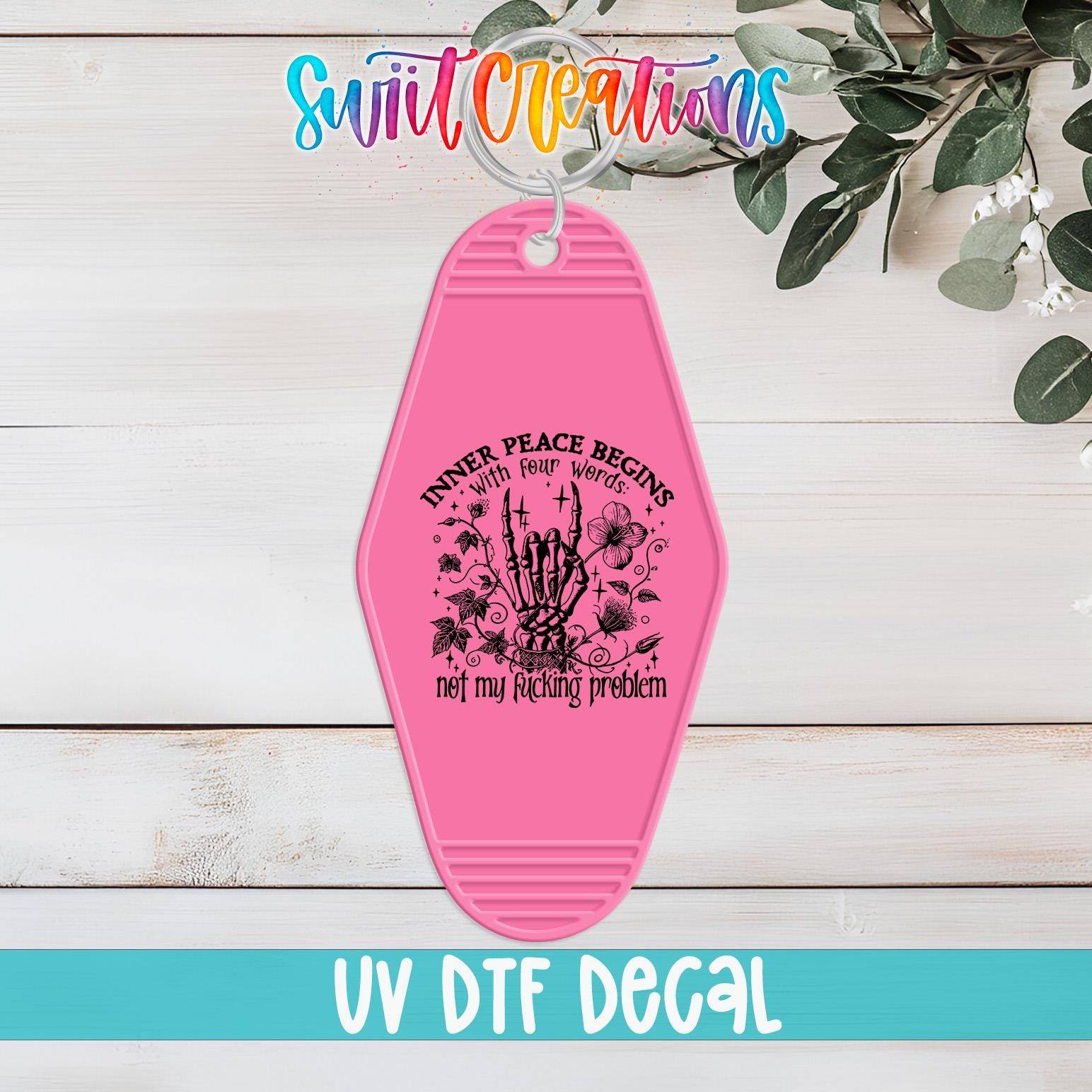 A pink keychain with a graphic design and text, placed on a wooden surface with greenery in the background.