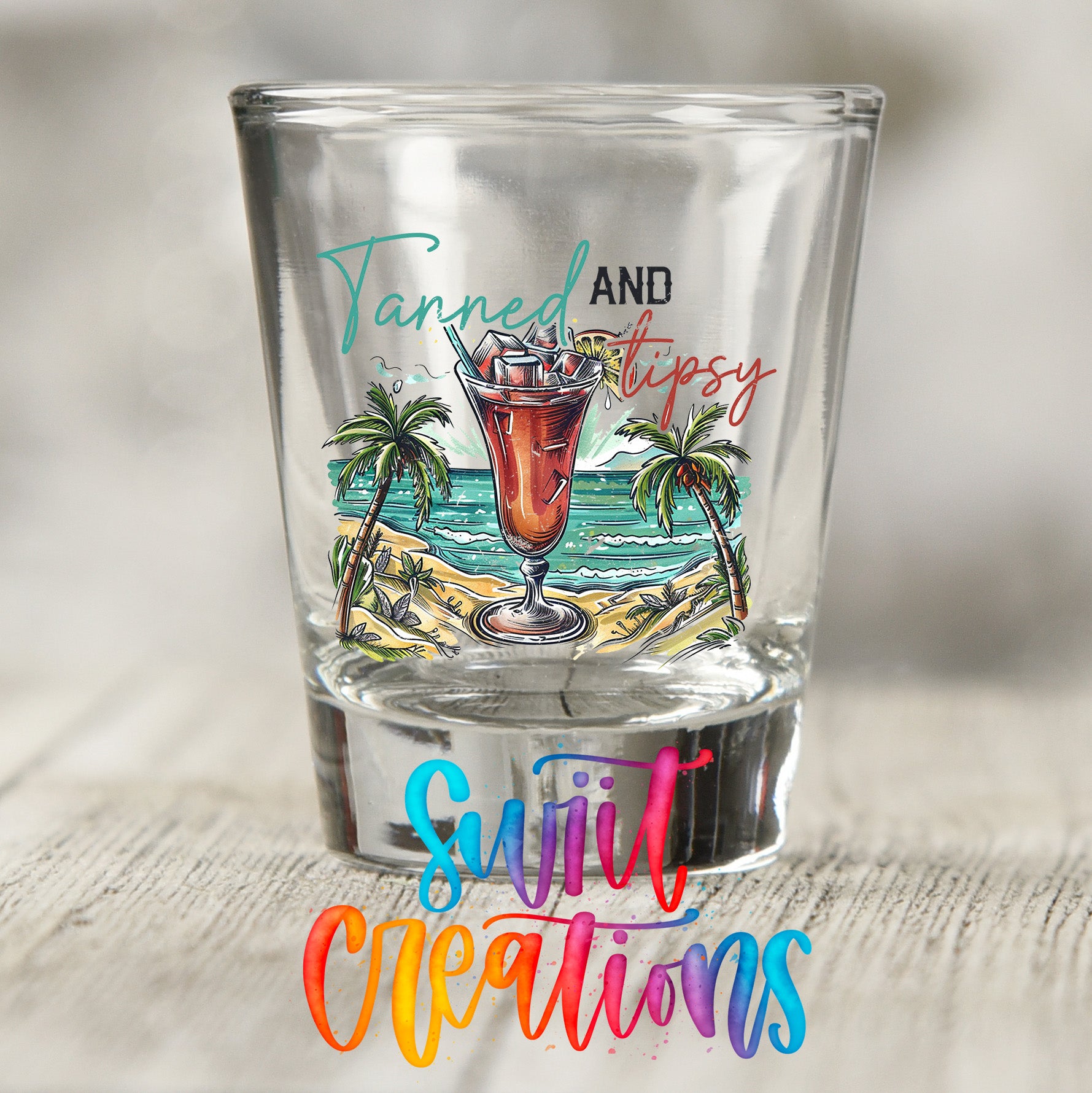 A shot glass with a tropical beach scene and a cocktail illustration, along with the text "Tanned and tipsy" and "Sweet Creations" written on it.