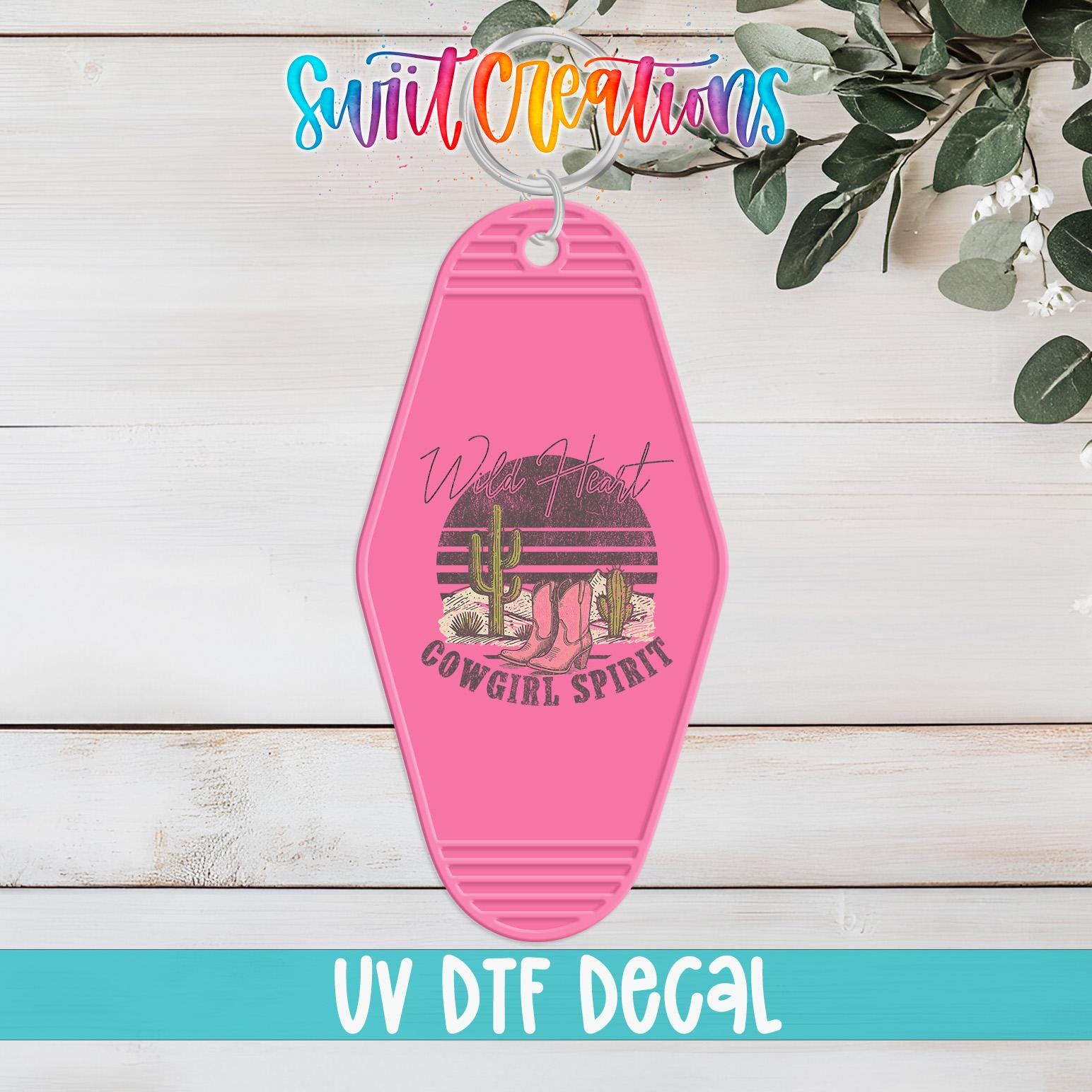A pink keychain with a graphic design featuring a cactus and the text "Wild West Cowgirl Spirit" is displayed on a wooden surface.