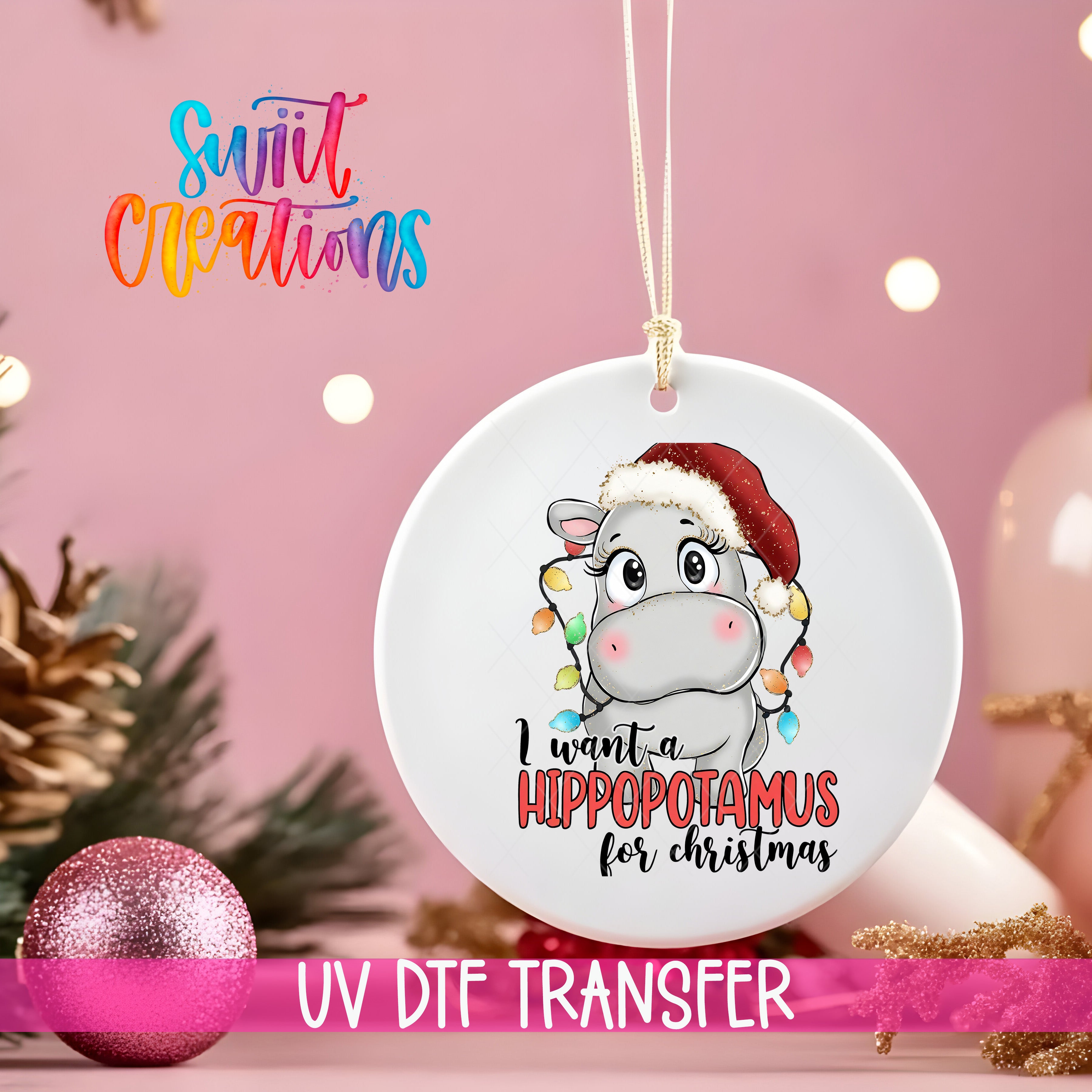 A white ceramic ornament with a cartoon image of a hippopotamus wearing a Santa hat, along with the text "I want a Hippopotamus for Christmas". The ornament is hanging from a string, and there are Christmas decorations in the background, including a pink ornament and a pine cone.